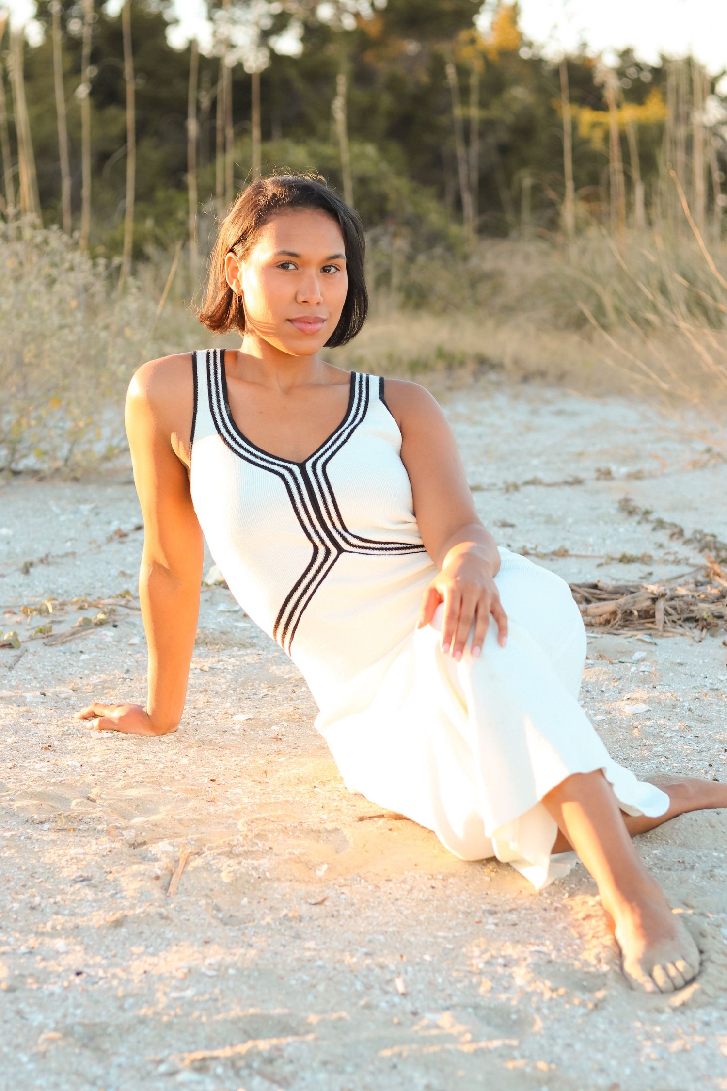 Woman in white dress with black trim sits barefoot on sandy beach at golden hour with greenery background.