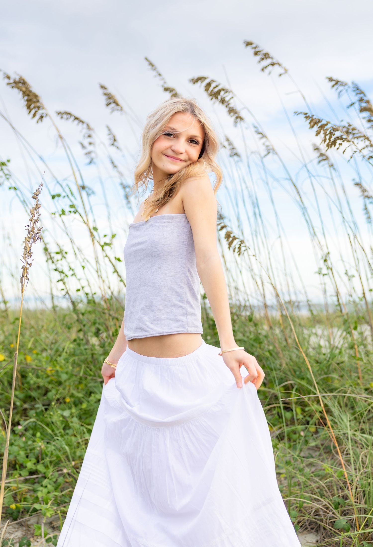 Smiling blonde woman in a gray tank top and flowing white maxi skirt poses among tall sea oats on a sunny coastal dune.