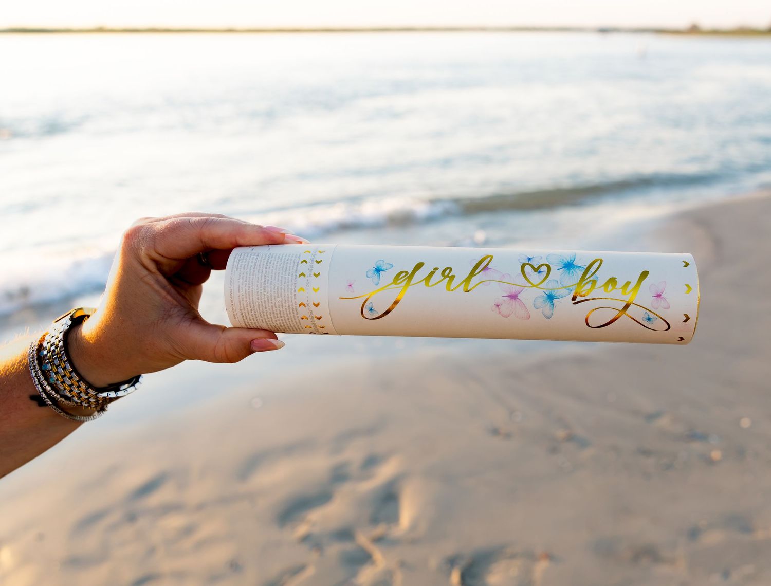 Hand holding a gender reveal tube with girl or boy text on a beach at sunset.