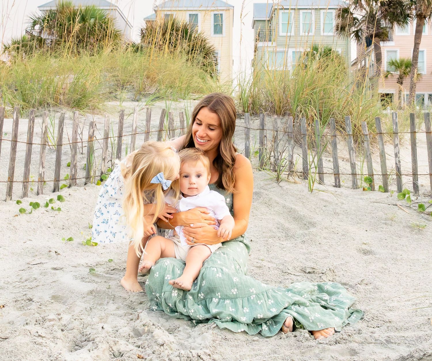 A smiling mother in a green floral skirt sits on a sandy beach with a young girl and a baby, with dune grass and houses behind.