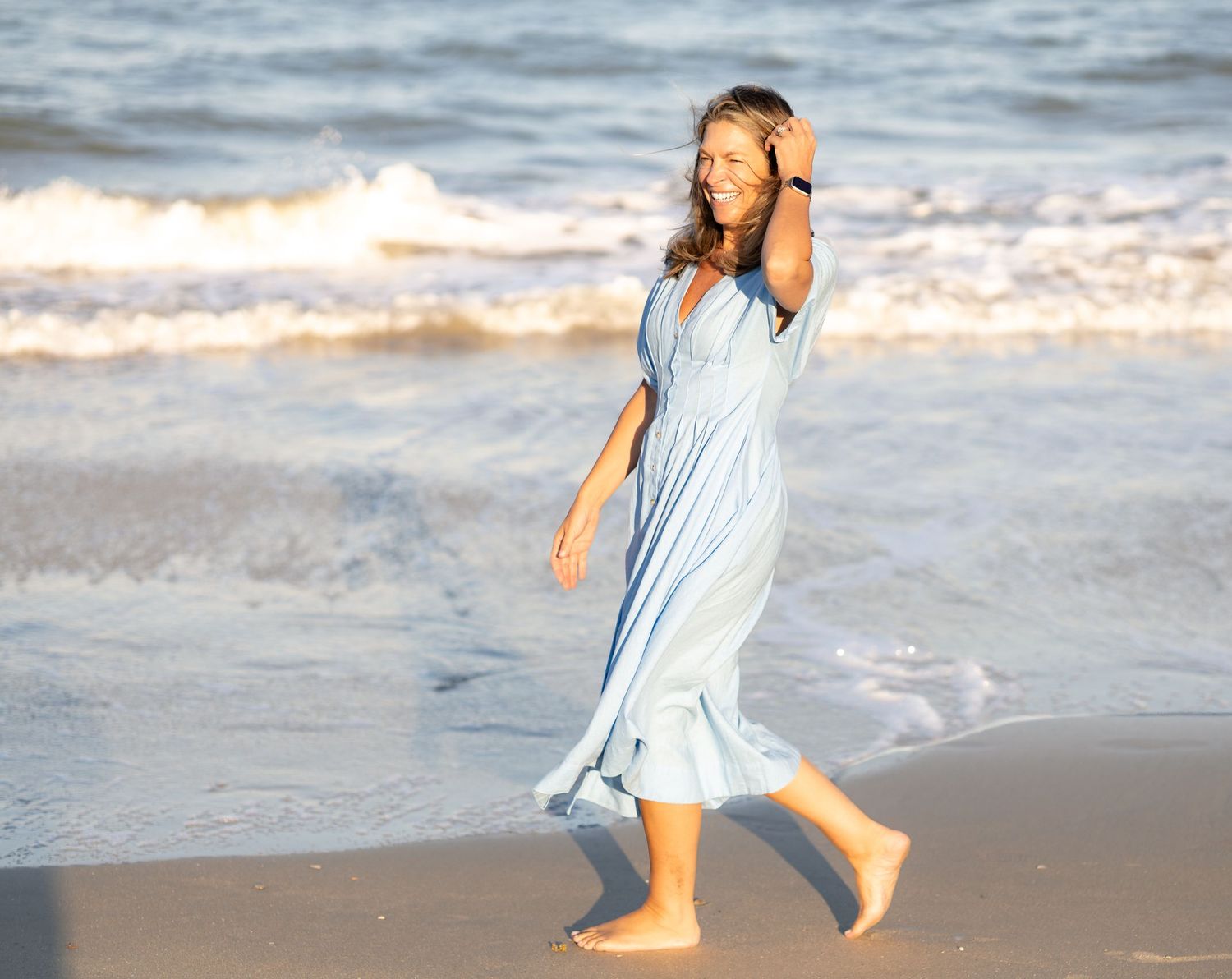 Woman in light blue maxi dress walking barefoot on sandy beach with ocean waves in background.