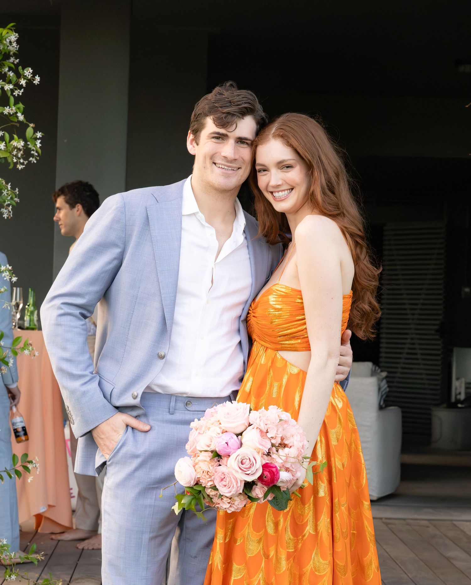 Couple smiling at wedding event, woman in orange dress holding pink bouquet.