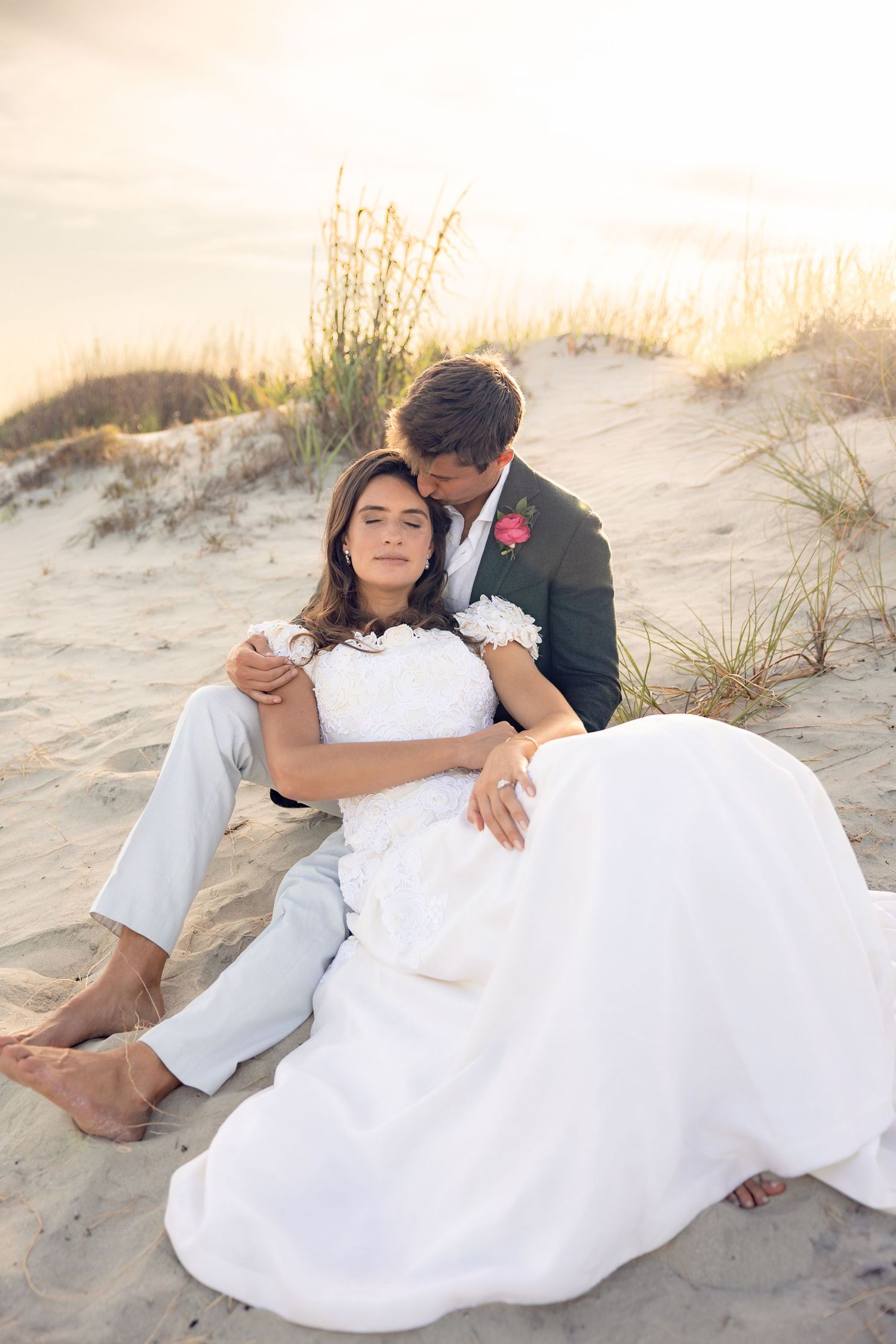 Bride and groom sitting together on sandy beach dunes at golden sunset.