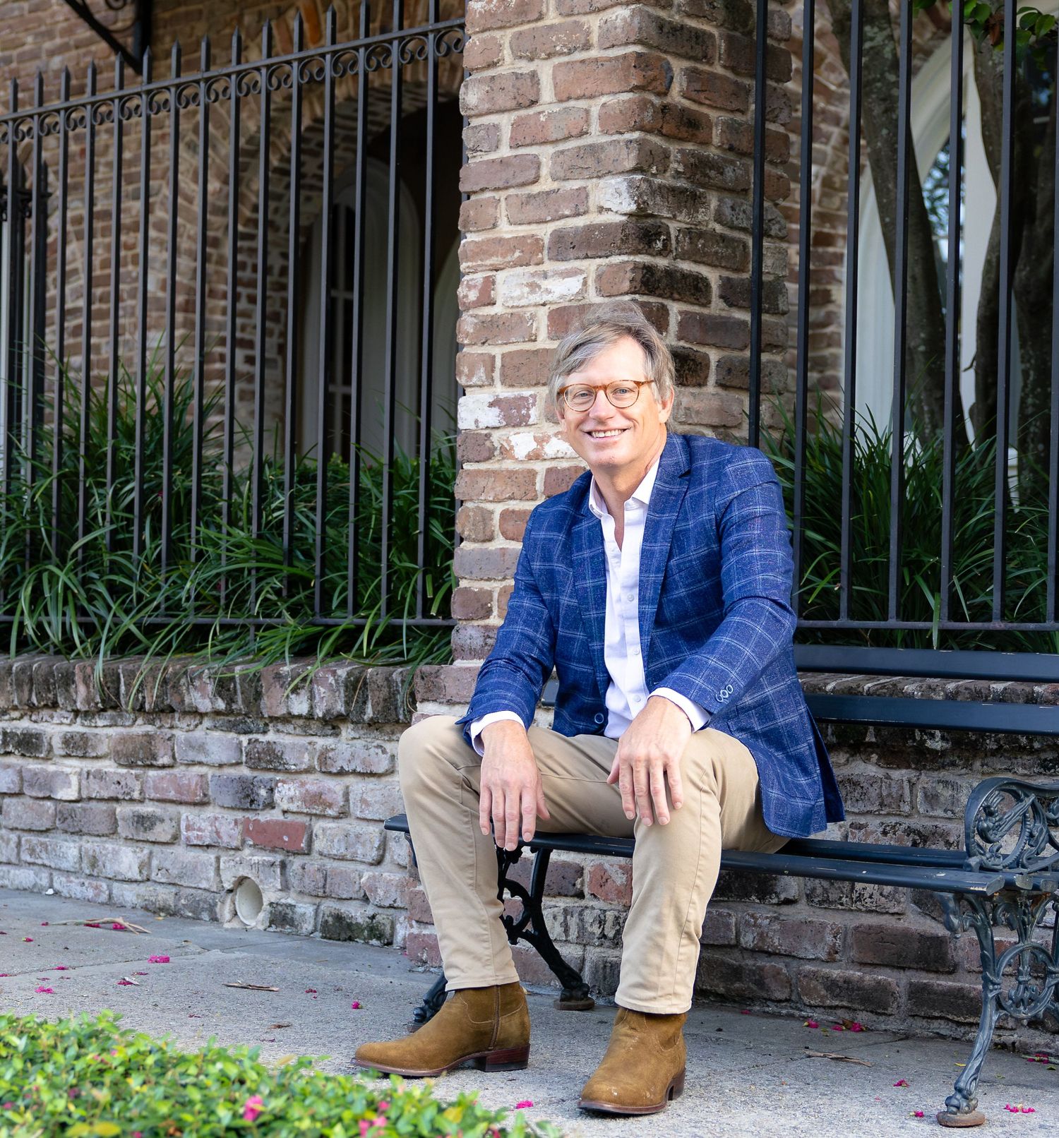 Man in blue plaid blazer sitting on bench outside brick building with iron fence.