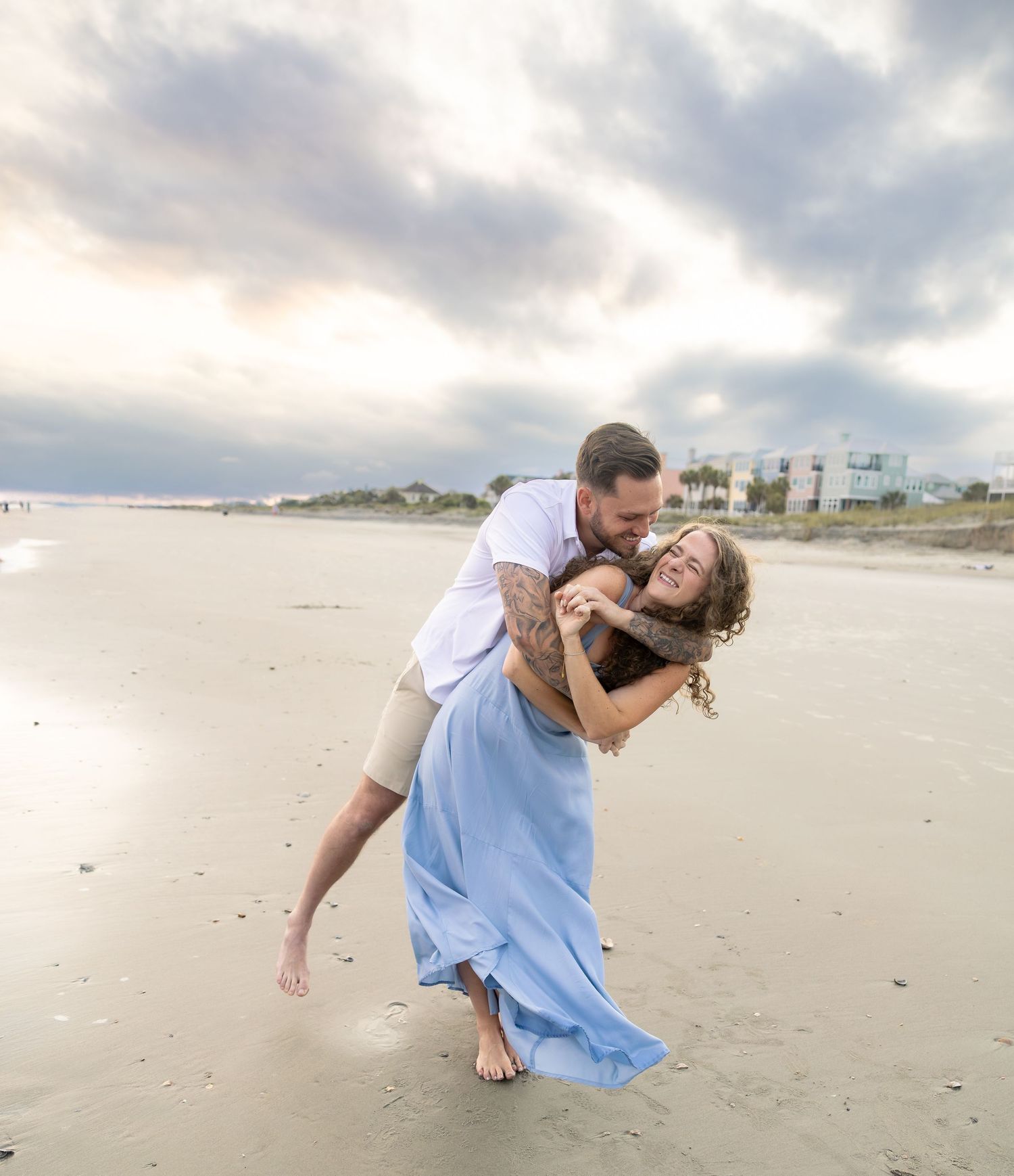 Couple shares romantic dip kiss on sandy beach at sunset during engagement photo shoot.