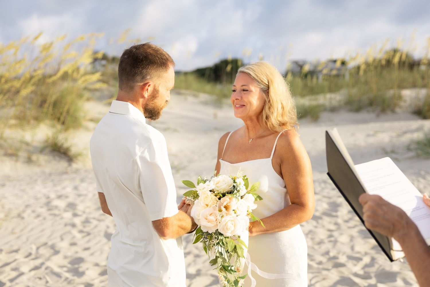 Couple exchanging vows on a sandy beach wedding, bride holding white bouquet, officiant reading ceremony.