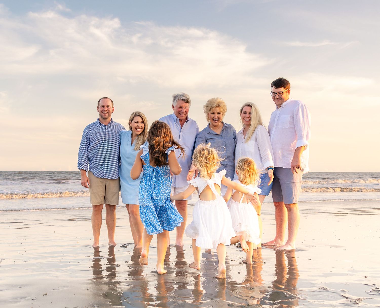 Multi-generational family posing together on a sunny beach with ocean waves, wearing coordinated blue and white outfits.