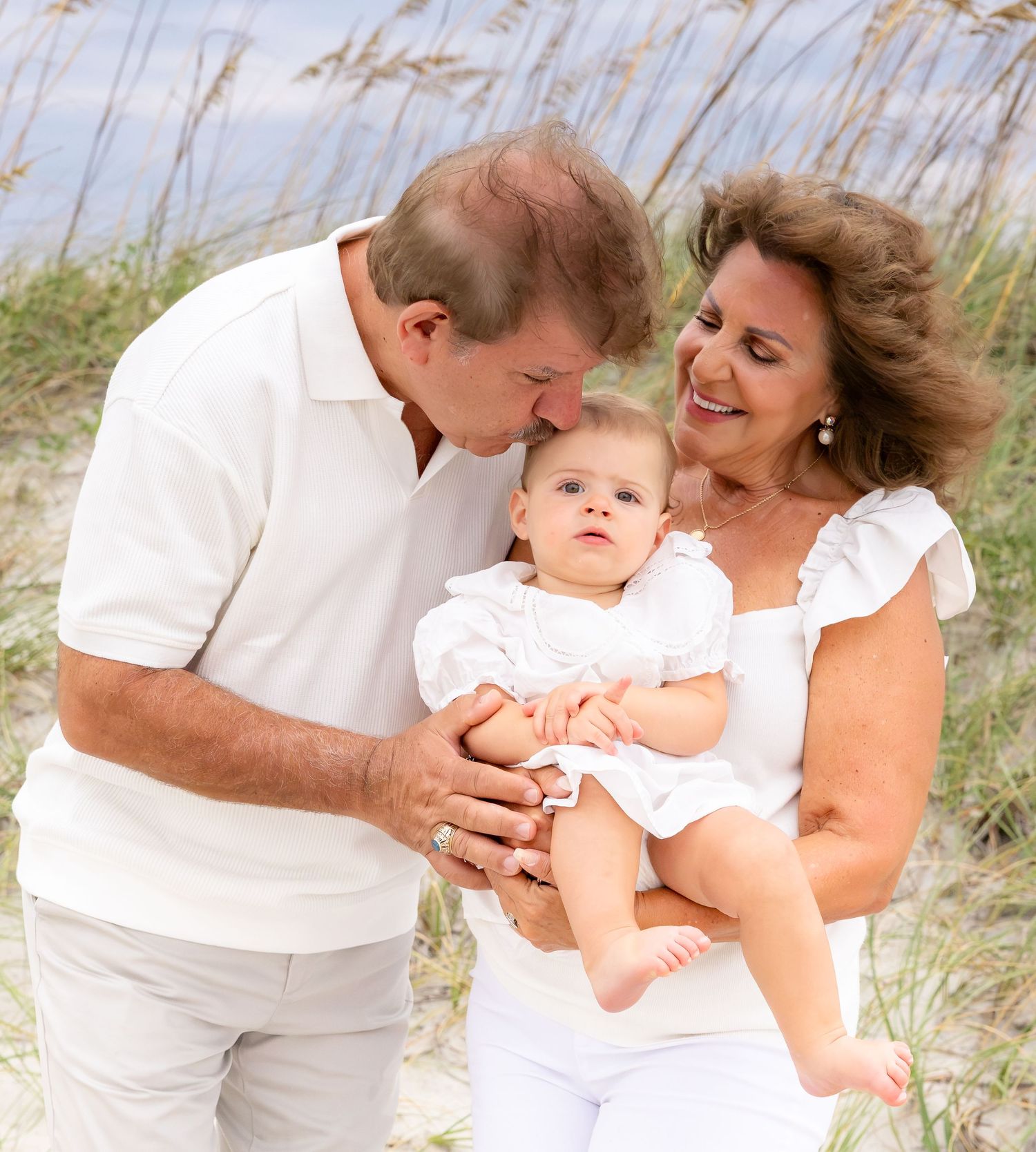 Grandparents holding baby on beach, smiling family in white outfits with sand dunes backdrop.