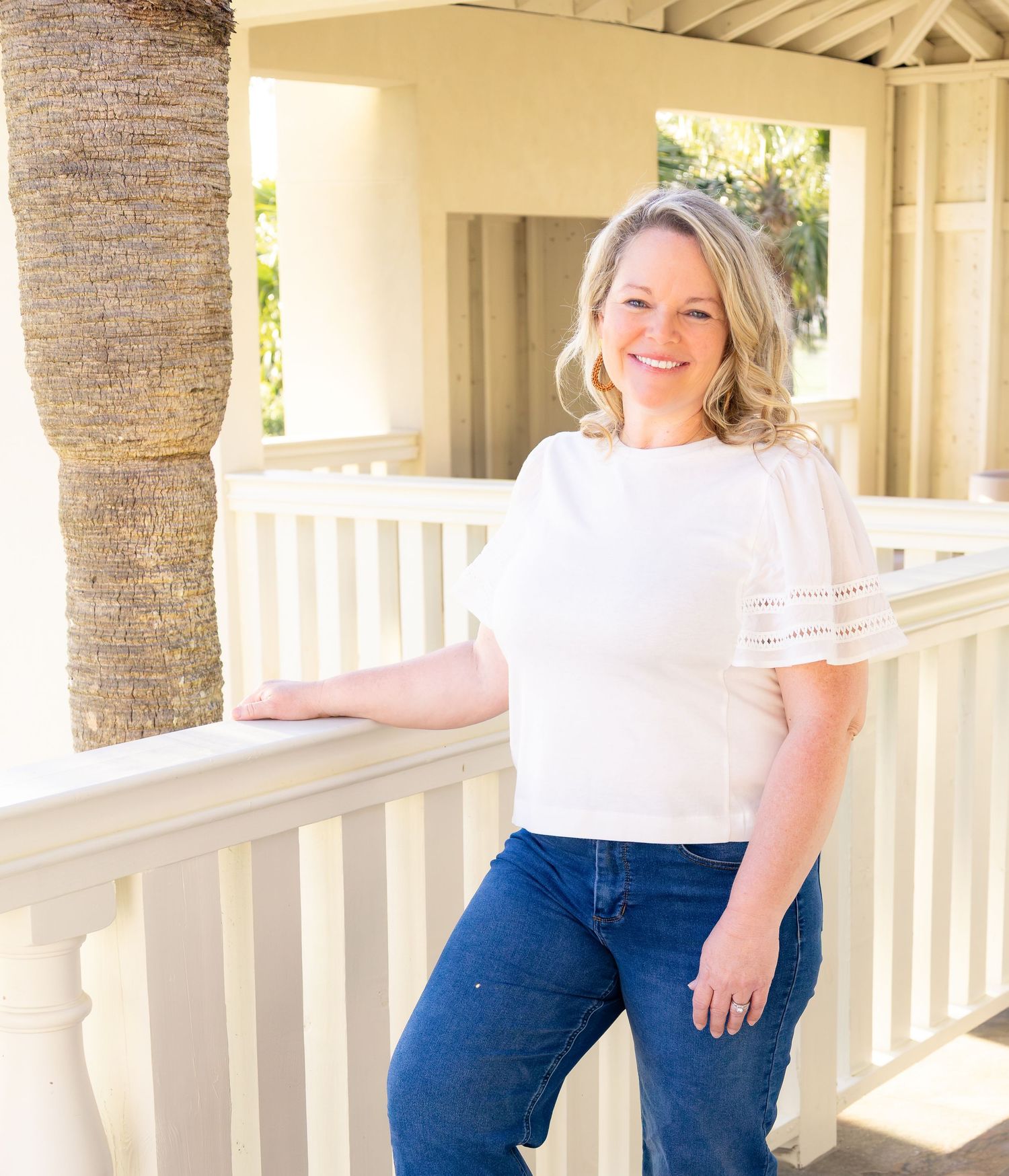 Smiling blonde woman in white top and jeans leaning on white porch railing of a tropical home.