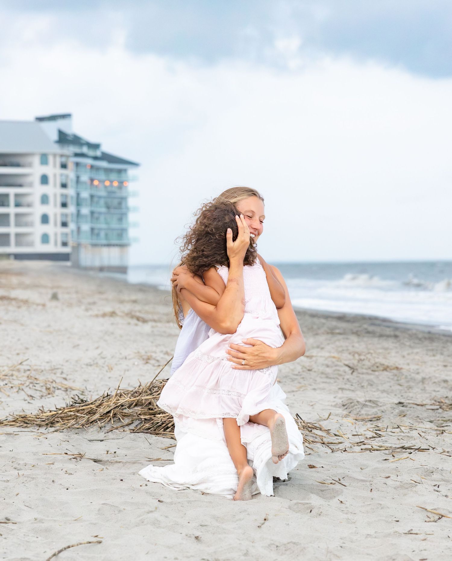 Woman in white dress kneeling on beach, hugging child, with ocean and hotel in background.