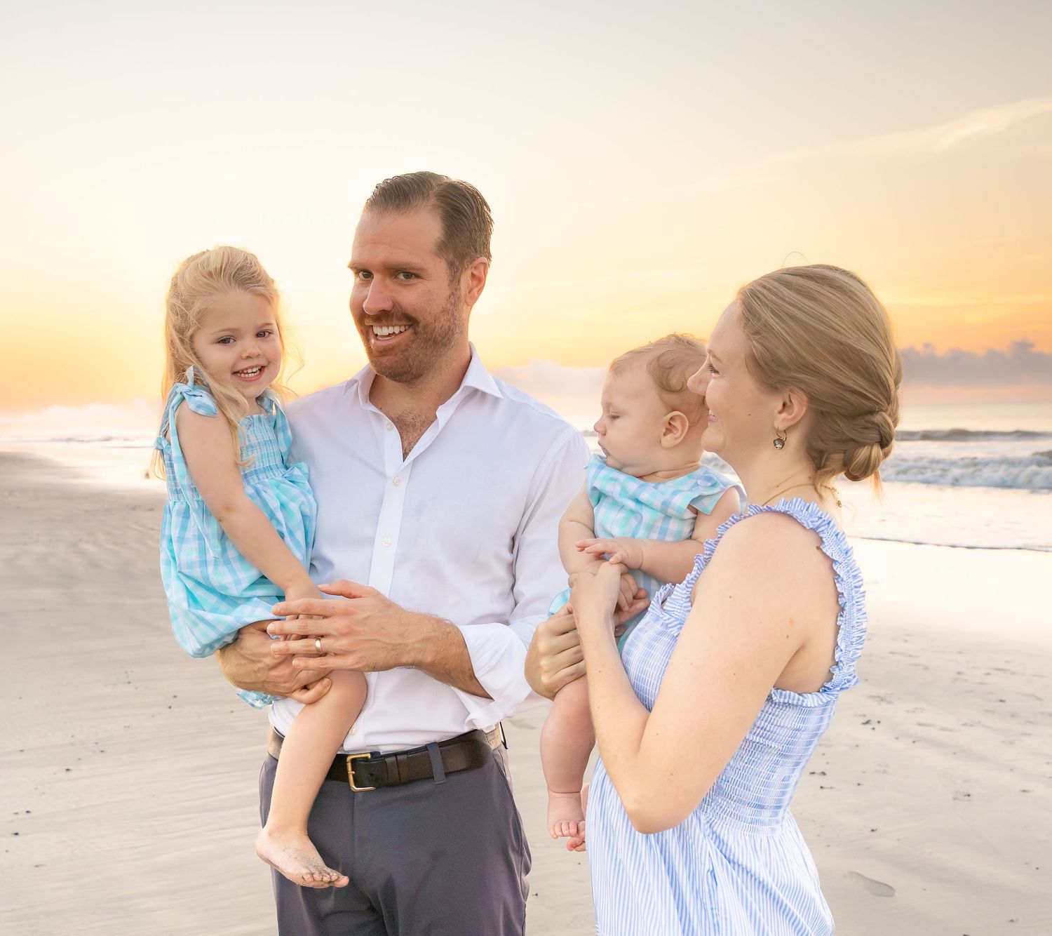 Family of four smiling on a beach at sunset, parents holding two young children in matching blue outfits.