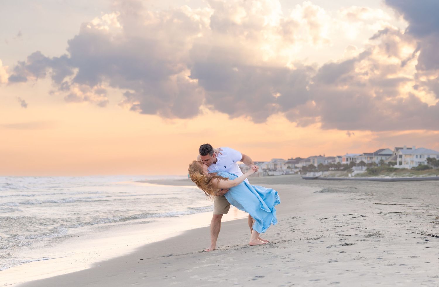 Couple sharing a romantic dip kiss on a sandy beach at sunset with pastel clouds and coastal town in background.