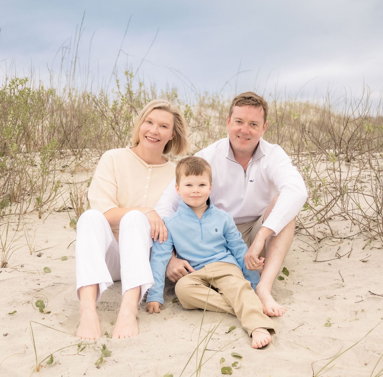 Family of three smiling together on sandy beach dunes, parents and young boy in casual coastal attire.