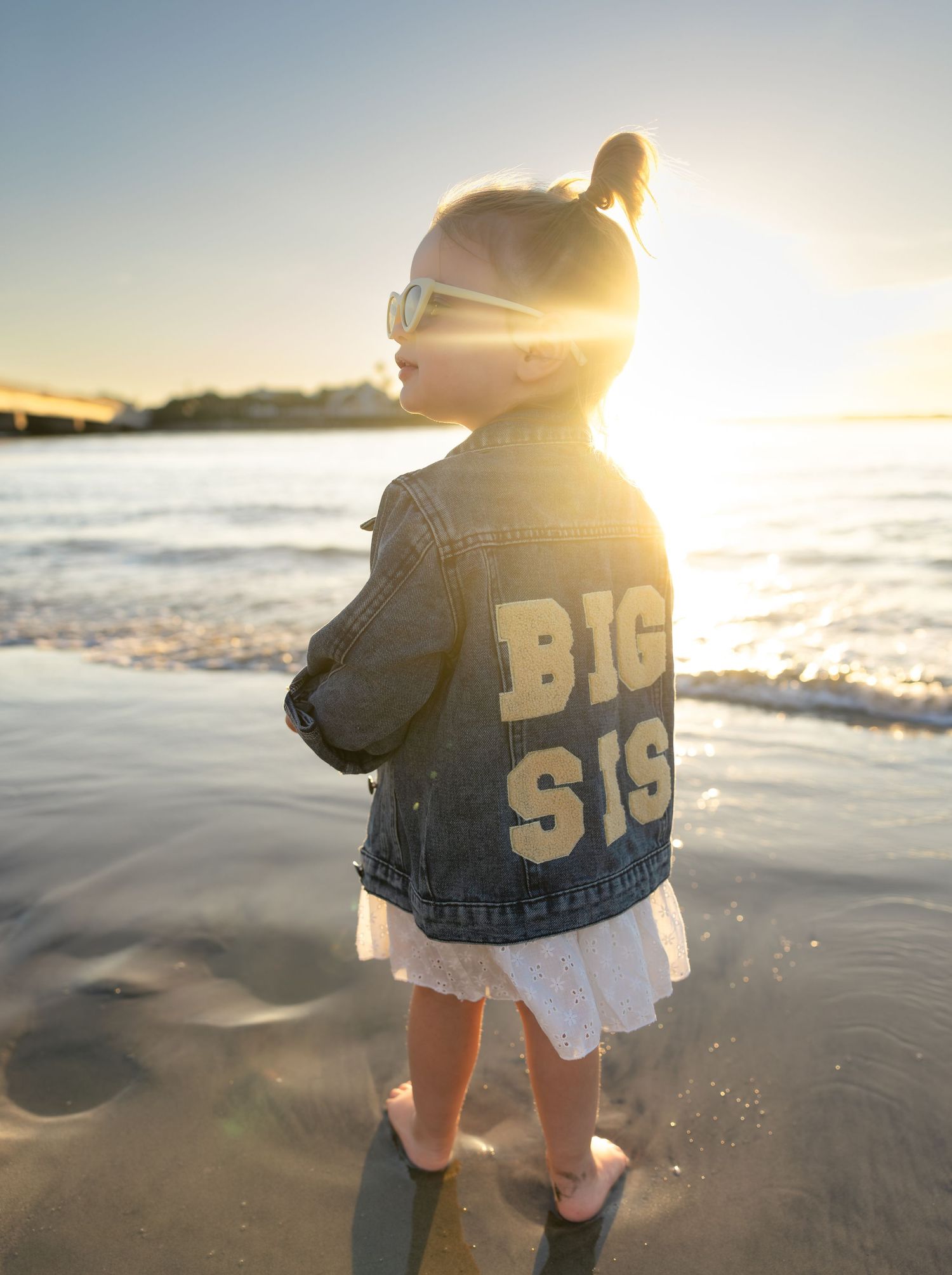 A toddler girl wearing a denim Big Sis jacket and sunglasses stands barefoot on a sunny beach at sunset.