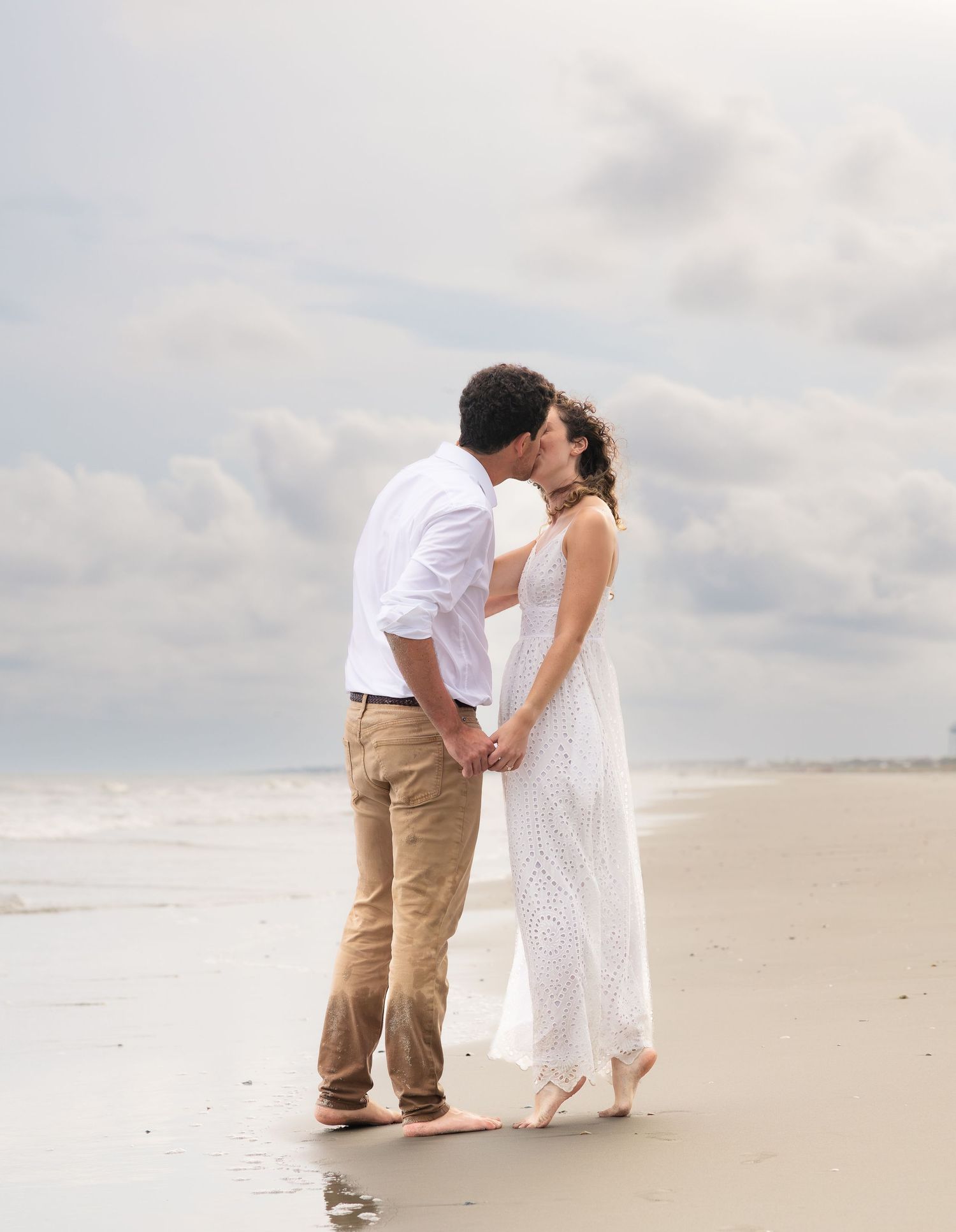 Couple kissing on a quiet beach at the water's edge, he in a white shirt and khakis, she in a flowing white lace dress, under a cloudy sky.