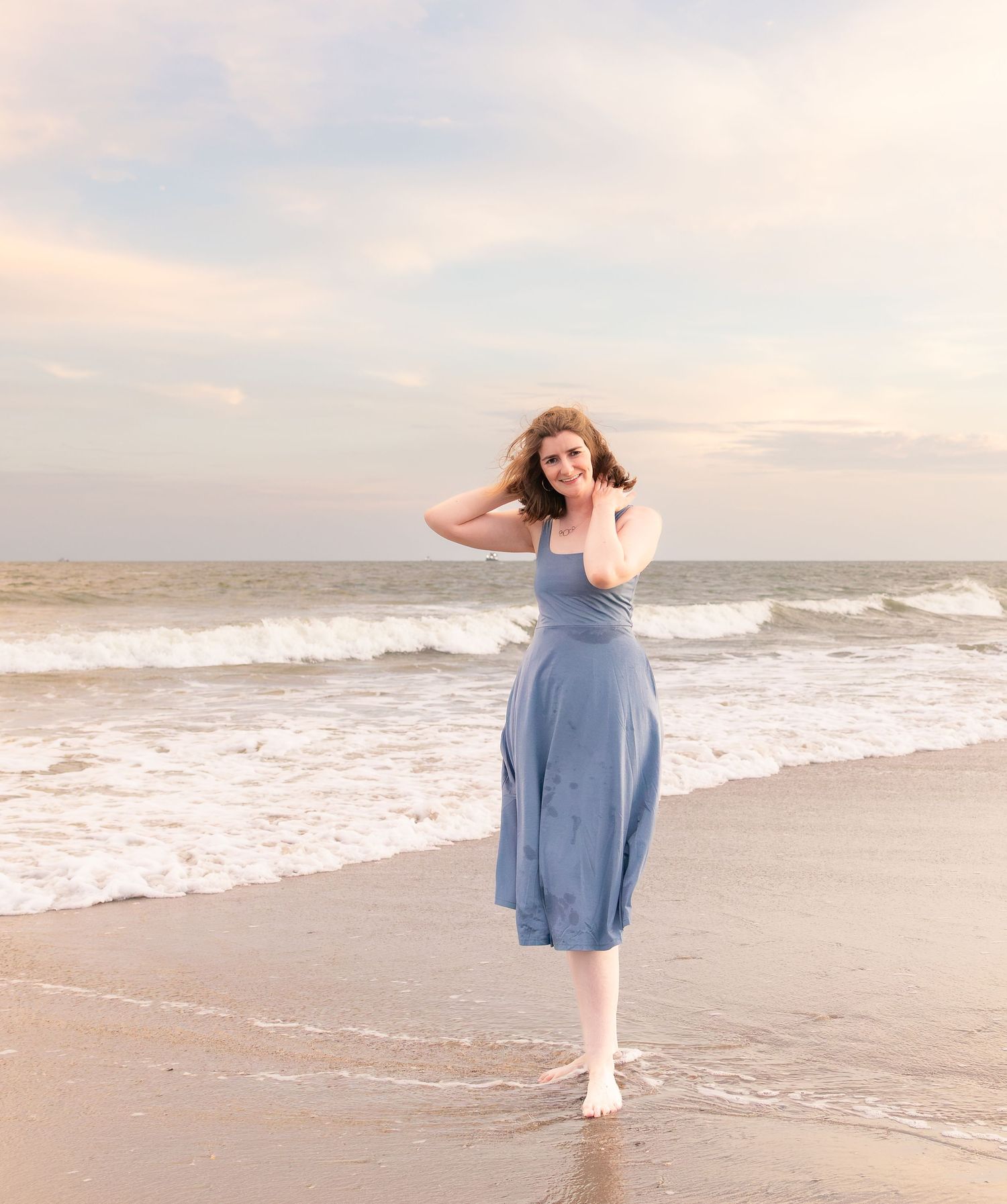 Woman in blue midi dress posing barefoot on sandy beach with ocean waves and pastel sunset sky.