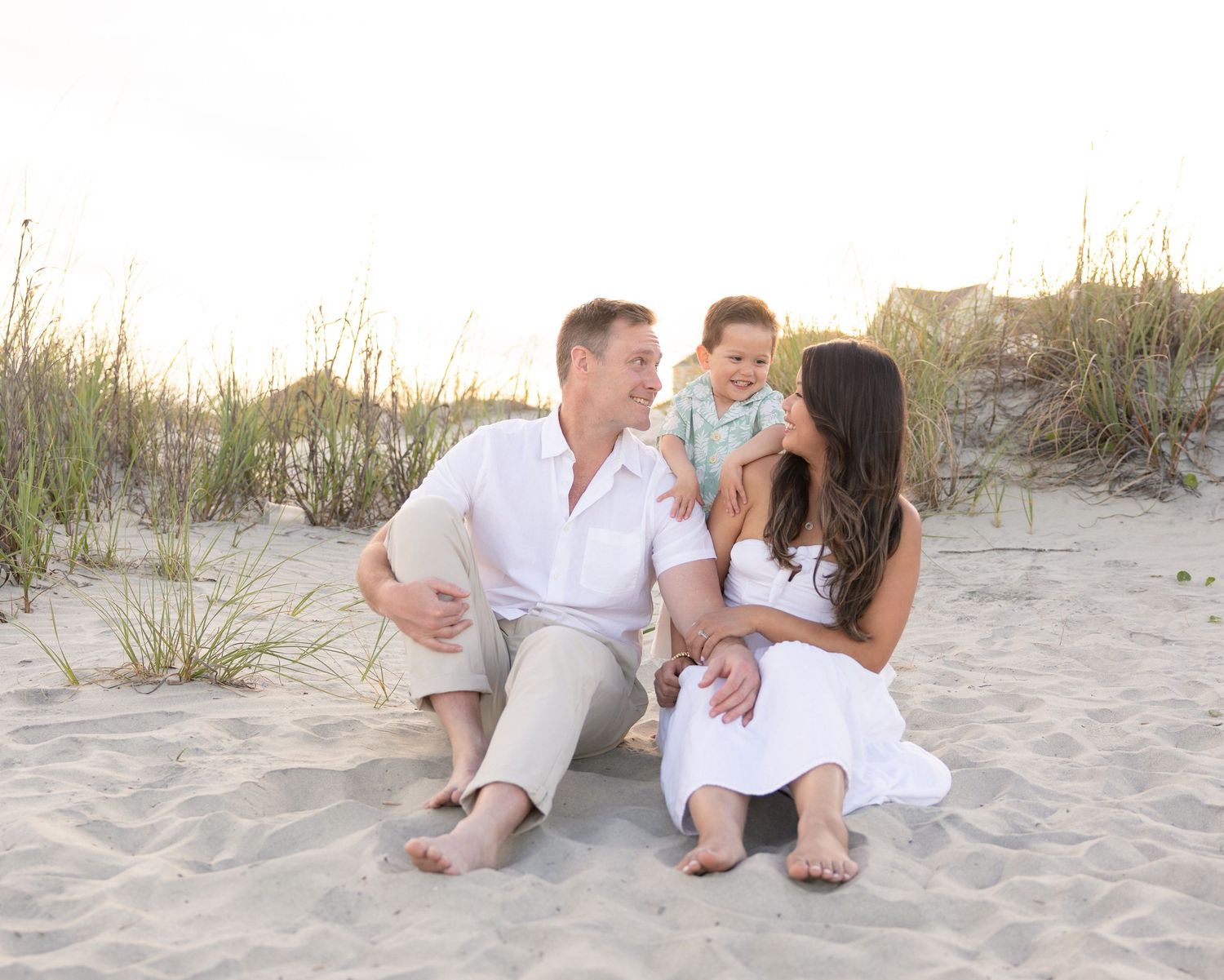 Family of three sitting on beach sand, parents smiling at toddler boy, dunes and grass in background.