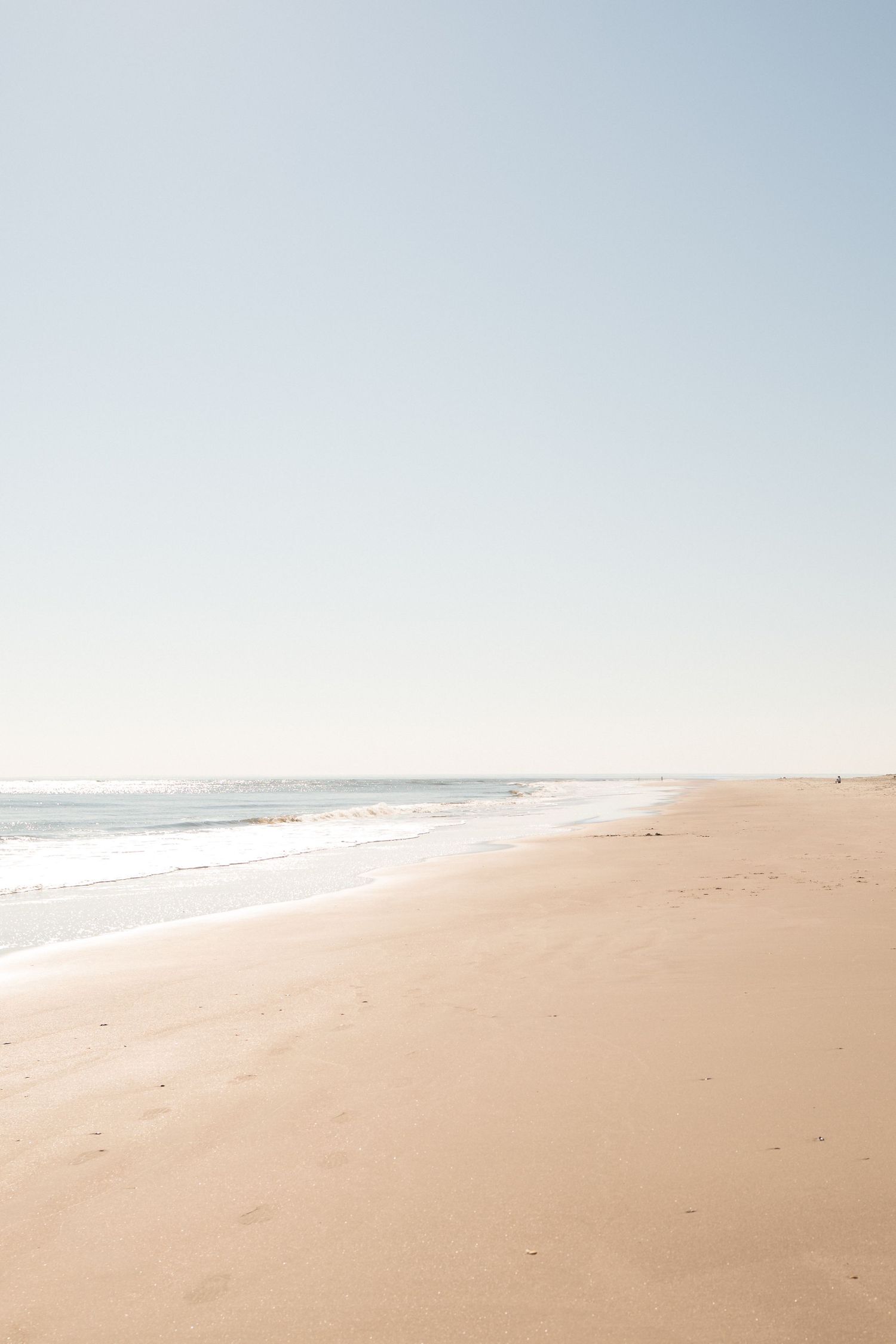 A serene, empty sandy beach stretches along calm ocean waves under a clear pale blue sky on a sunny day.