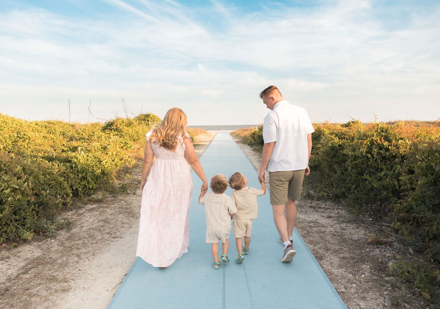 Family of four walking hand-in-hand down a scenic beach path surrounded by lush greenery under a blue sky.