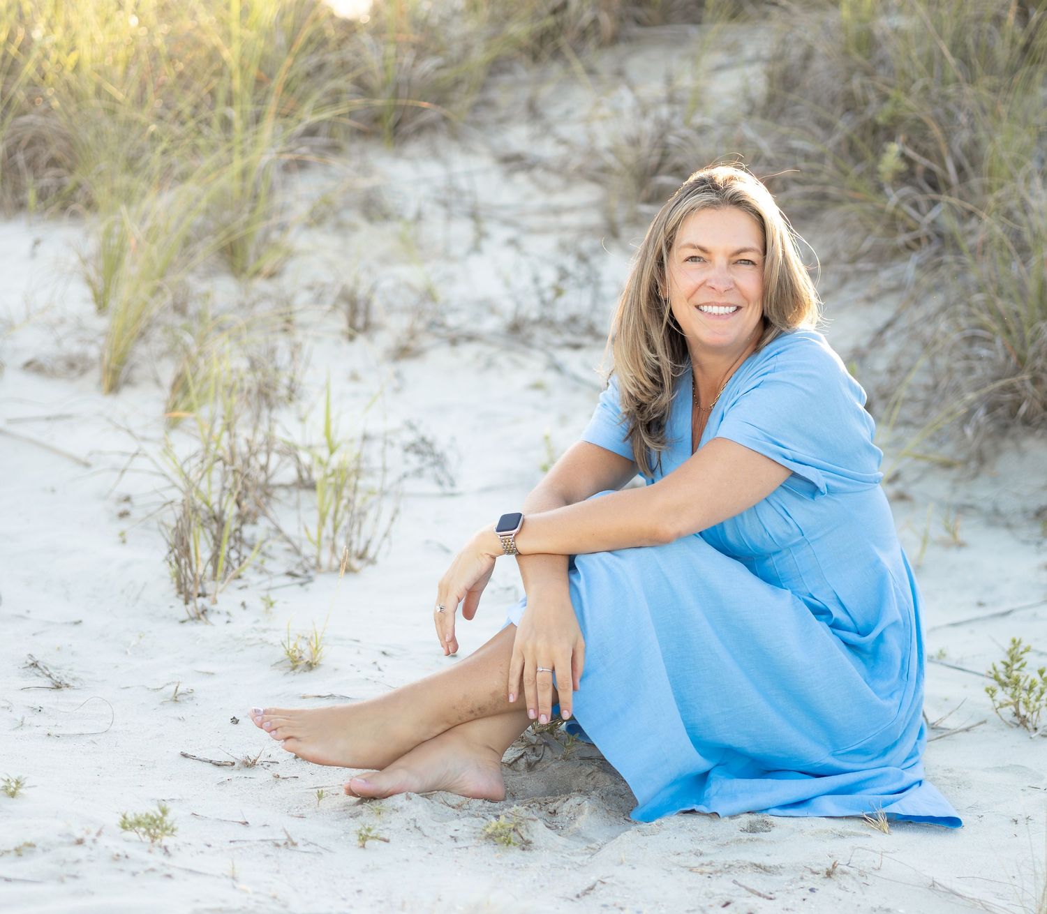 Woman in blue dress sitting barefoot on white sand beach dunes, smiling outdoors in natural light.