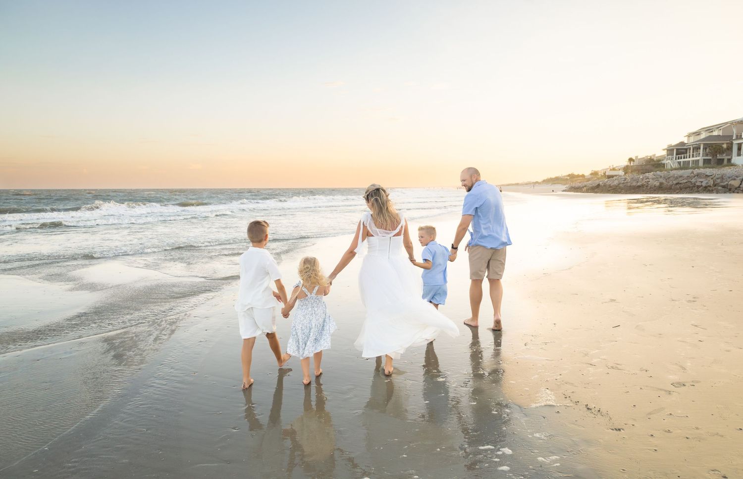 Family of five walking hand-in-hand on a sandy beach at sunset, wearing white and blue outfits.