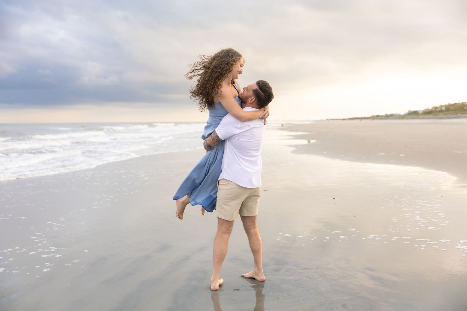 Couple embracing on beach at sunset, man lifting woman in blue dress near ocean waves.