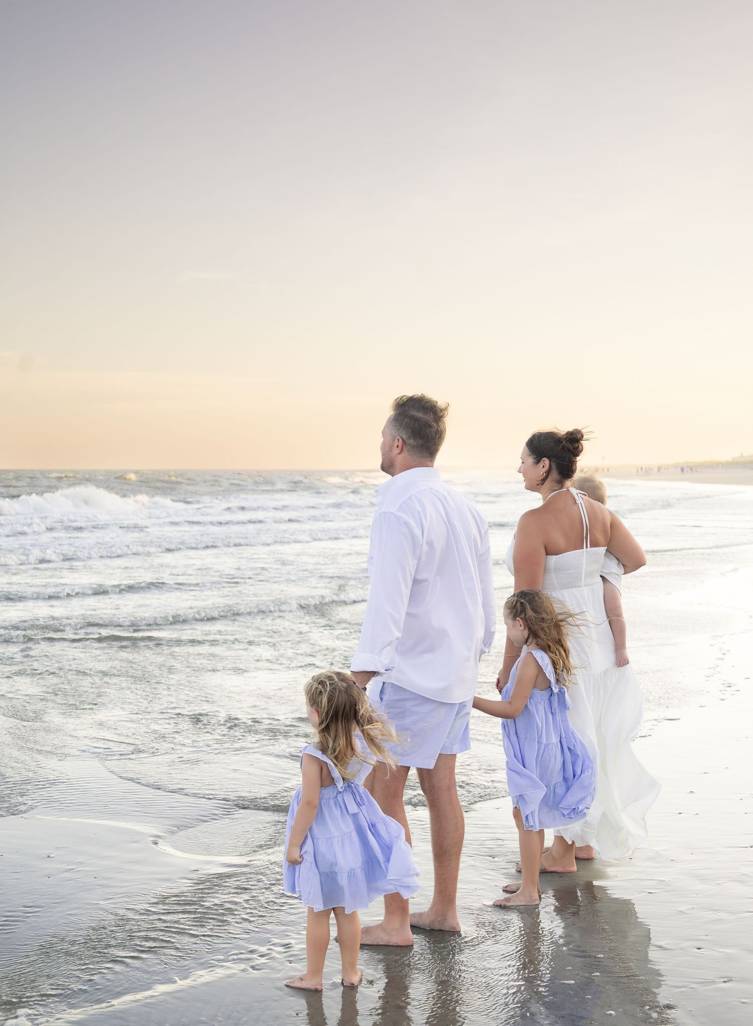 Family of four holding hands while watching ocean waves at sunset on a sandy beach.