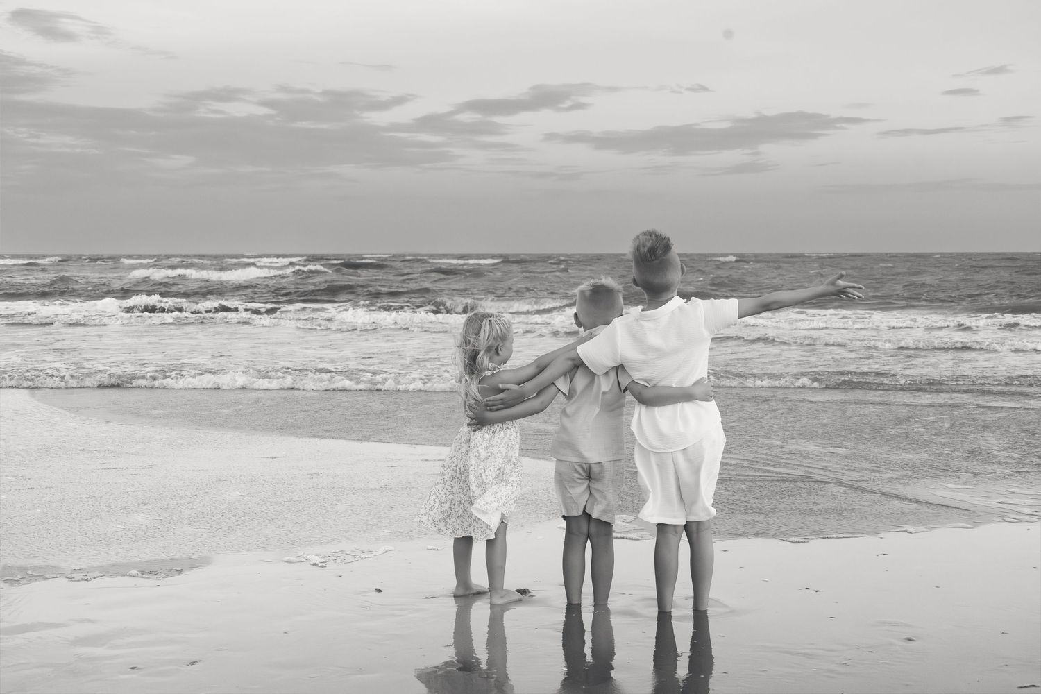 Black and white photo of three children hugging on a beach shore with ocean waves in the background.