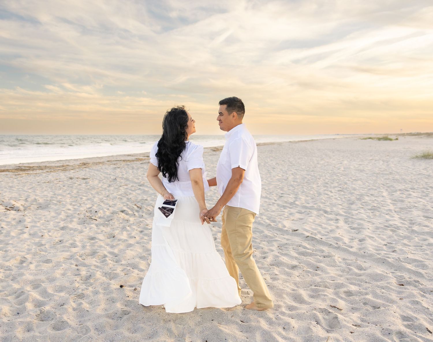 A couple holds hands on a sandy beach at sunset, woman in white dress, man in white shirt and khakis.