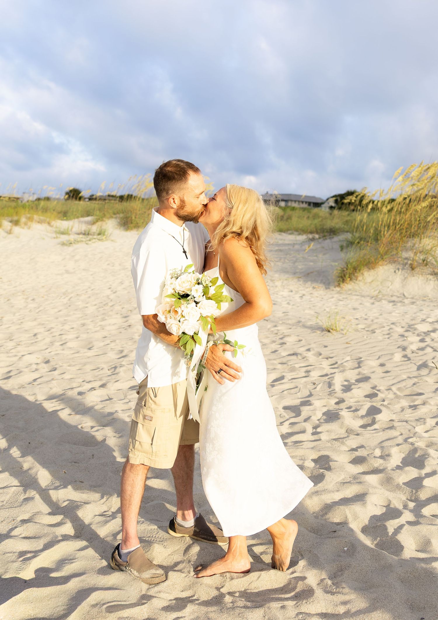 Couple kissing on sandy beach, bride in white dress holding bouquet, groom in white shirt and khaki shorts, wedding day.