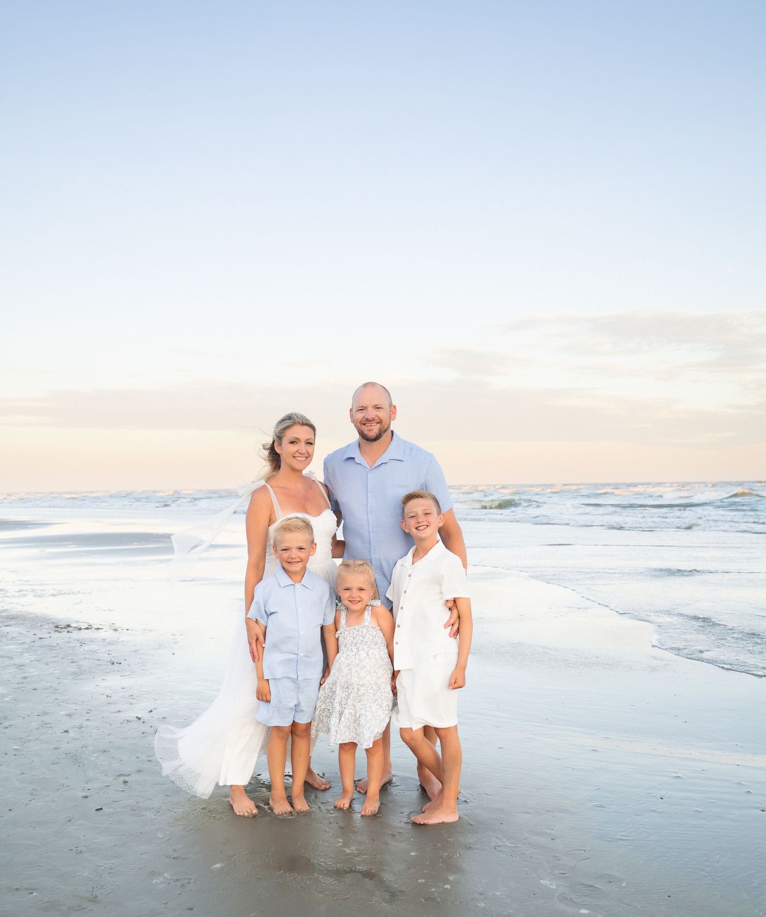 Family of five posing together on a sandy beach at sunset, wearing coordinated white and blue outfits.