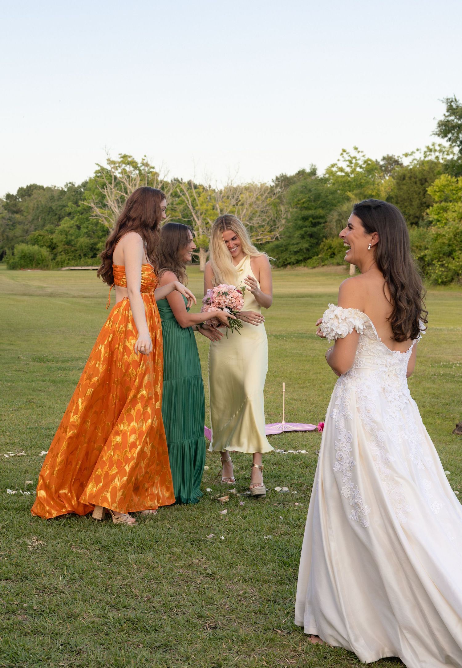 Bride in off-shoulder lace gown laughs with three bridesmaids in colorful dresses on green outdoor lawn.