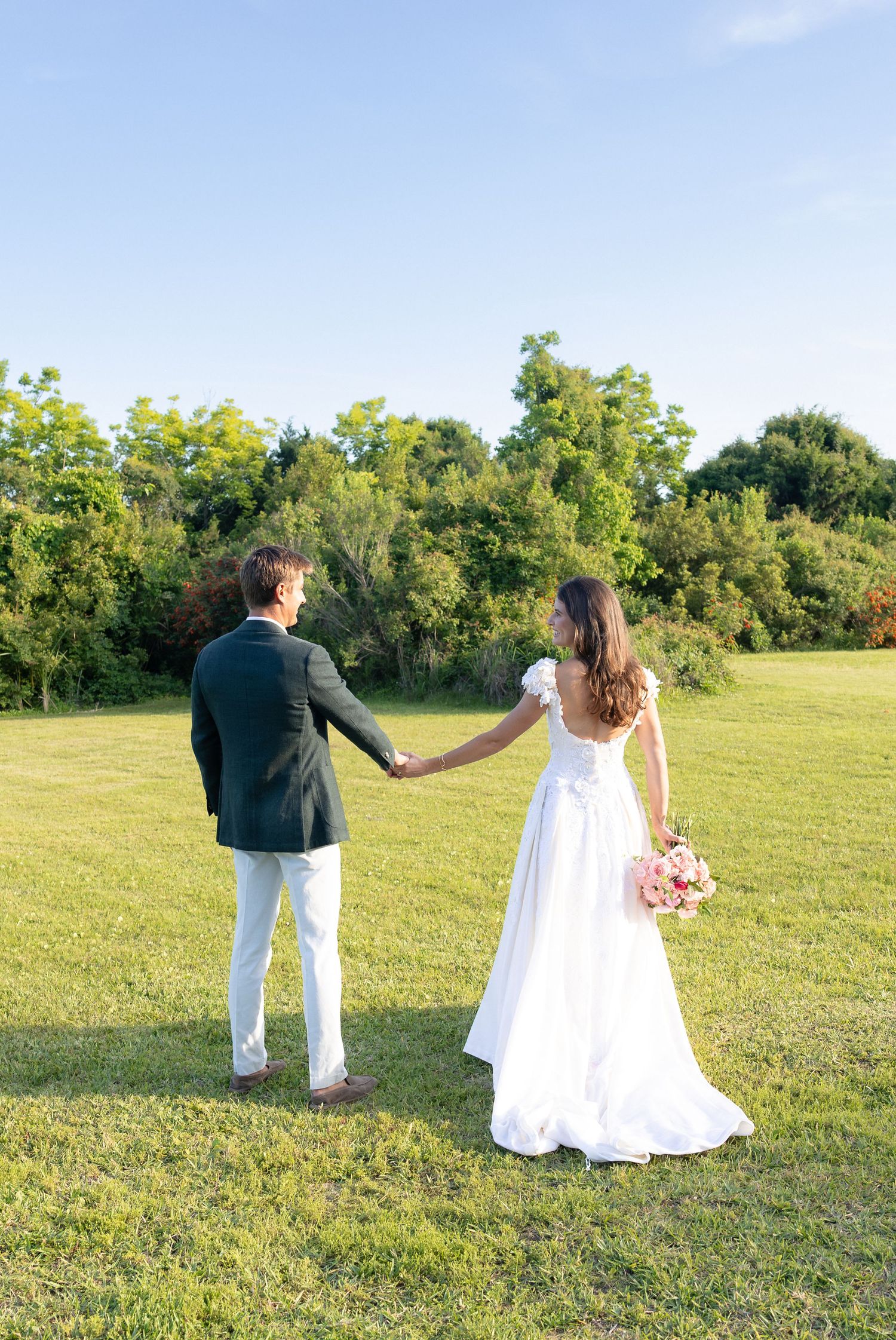 Bride and groom holding hands walking through lush green field on sunny wedding day.