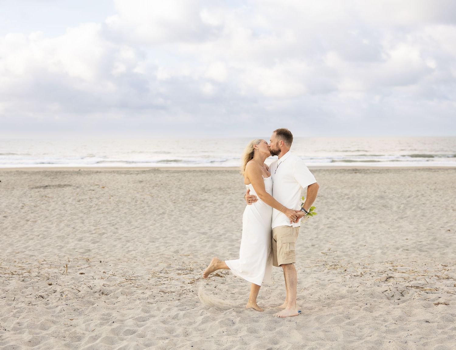 Couple sharing a romantic kiss on a sandy beach, dressed in white, with ocean waves and cloudy sky behind them.