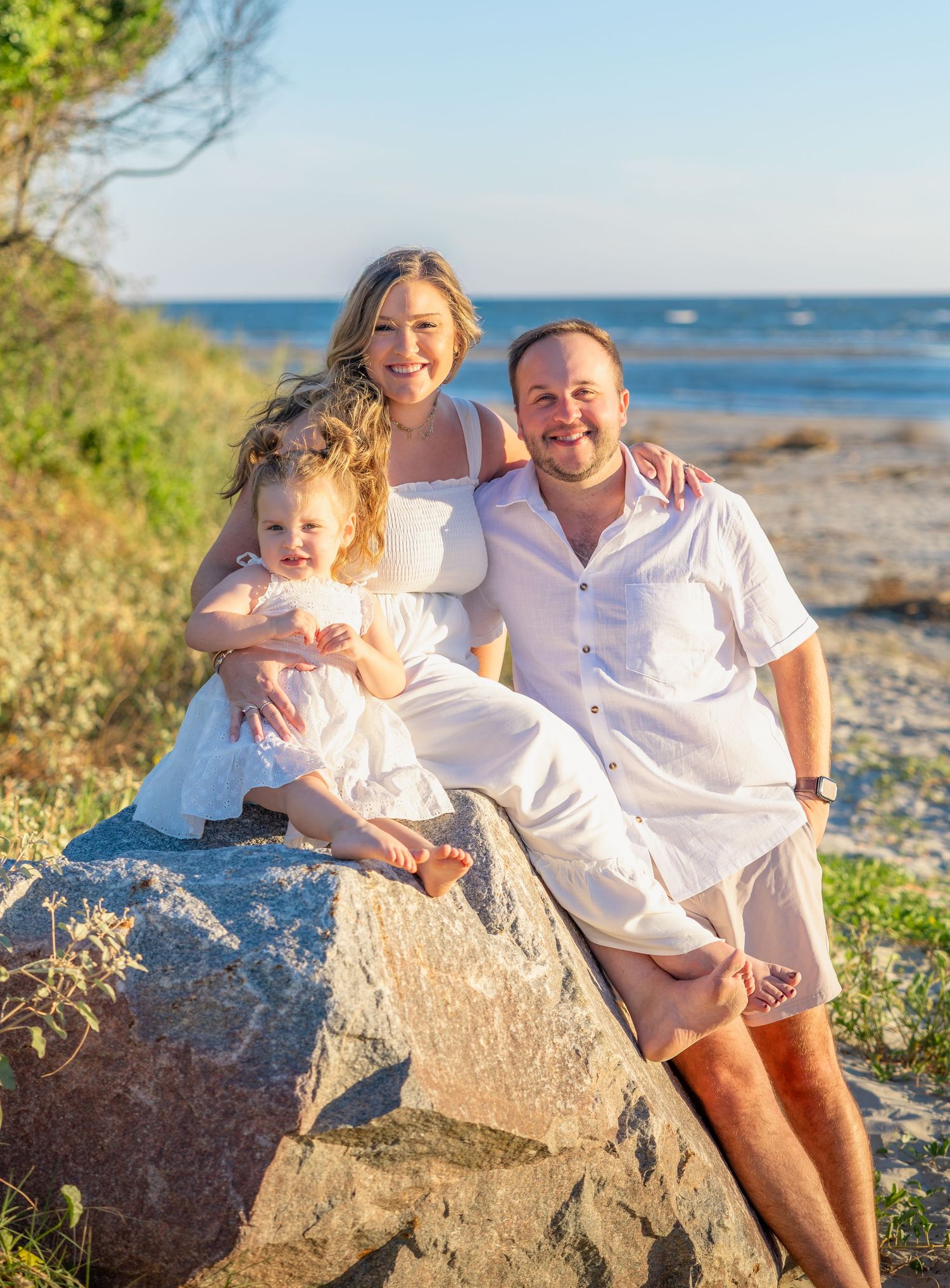 Smiling family of three in white outfits sit together on a large boulder with a toddler on mom's lap, with a sunny beach and ocean behind.
