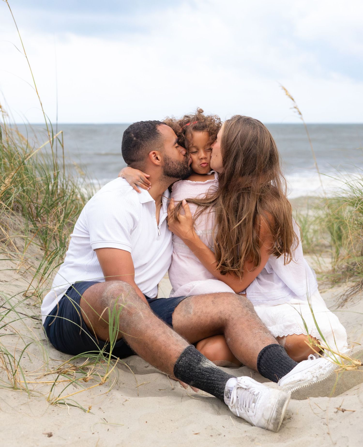 Family of three sitting on beach sand dunes, parents kissing toddler on cheeks, ocean in background.