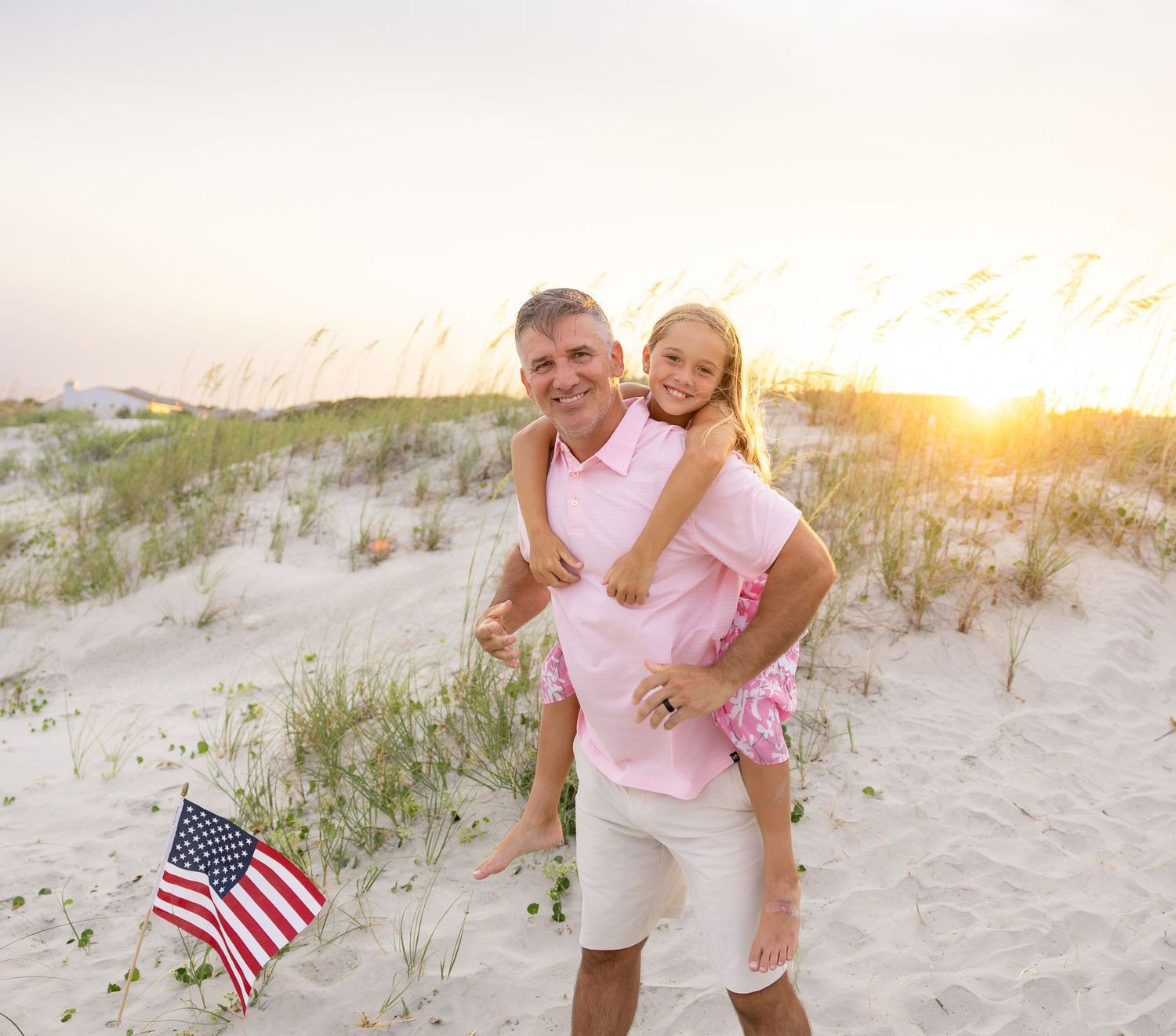 Man giving girl piggyback ride on sandy beach at sunset with American flag, both wearing pink shirts.