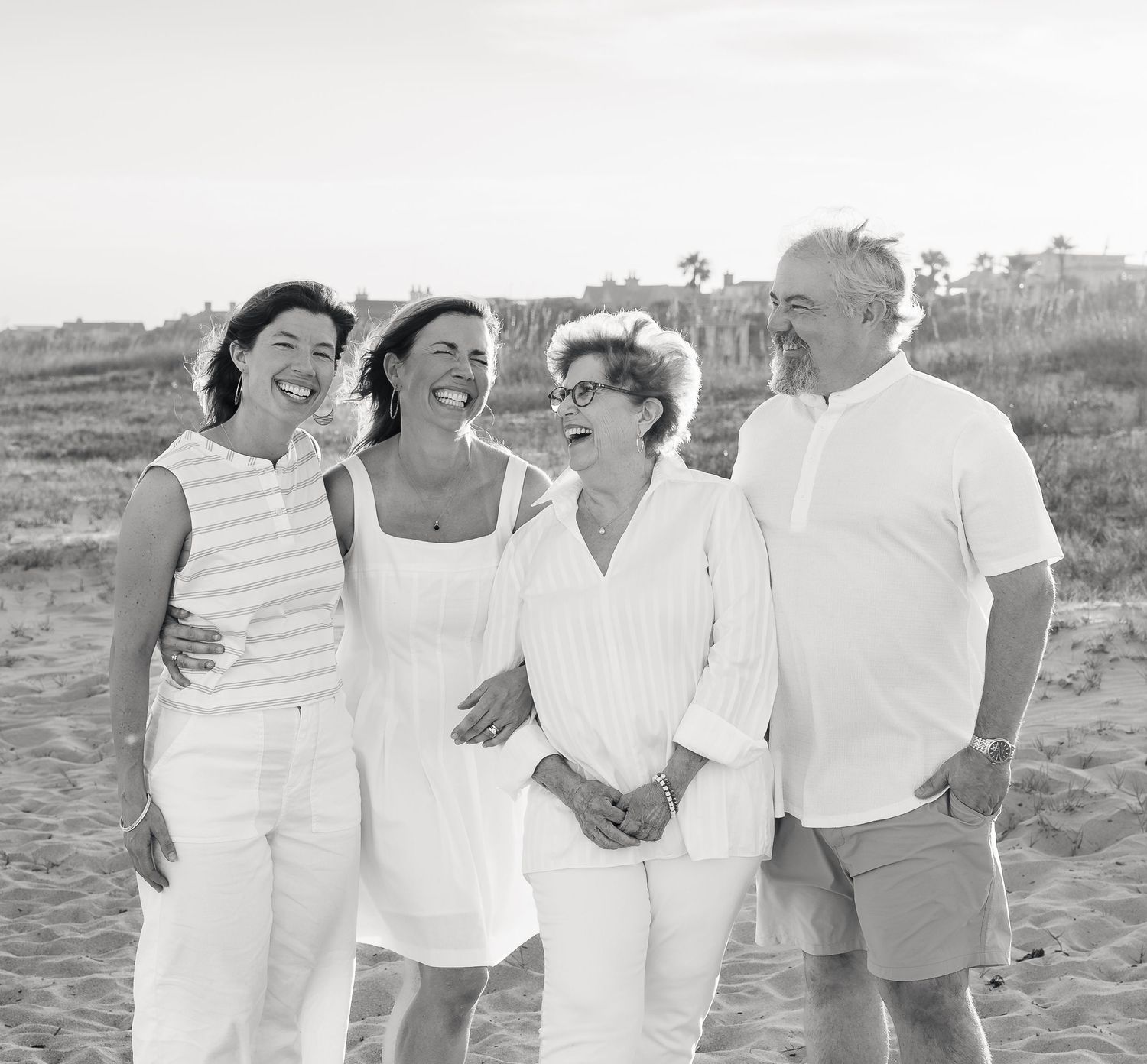 Black and white photo of four adults laughing together on a sandy beach, dressed in white summer outfits.