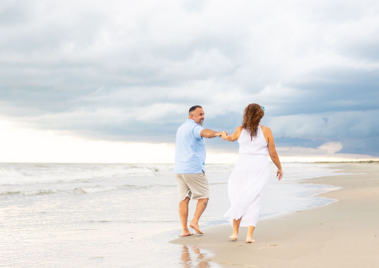 Couple holding hands and dancing on a sandy beach with dramatic cloudy skies and ocean waves behind them.