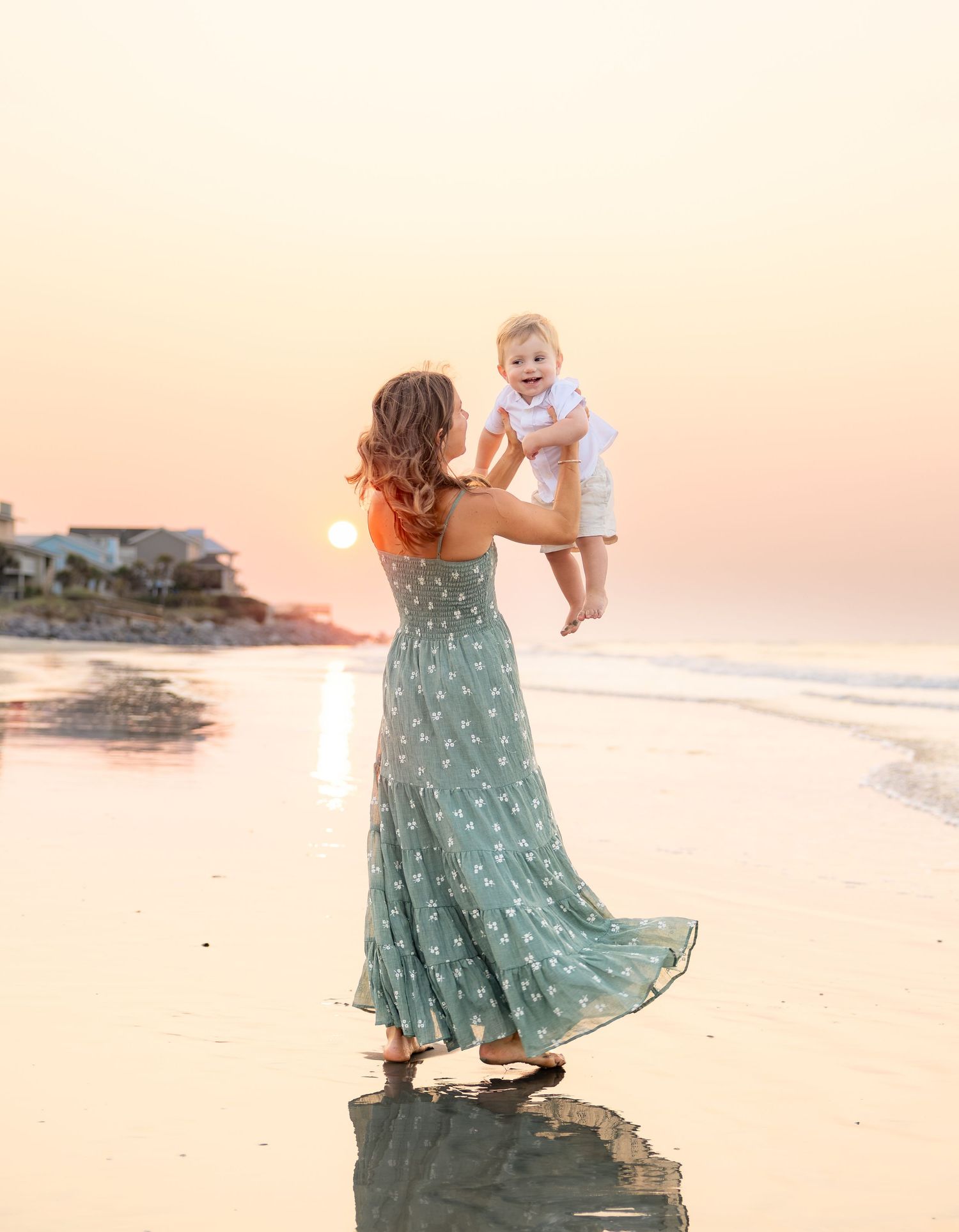 Mother lifting laughing baby in the air on a reflective beach during a golden sunset.