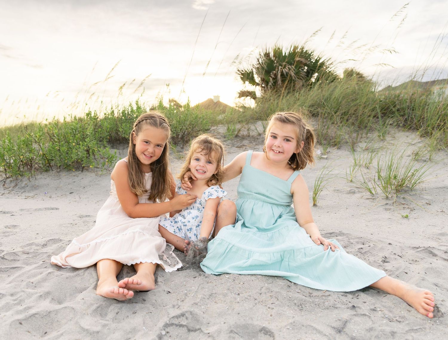 Three young girls in dresses smile together on a sandy beach at sunset with sea grass in the background.