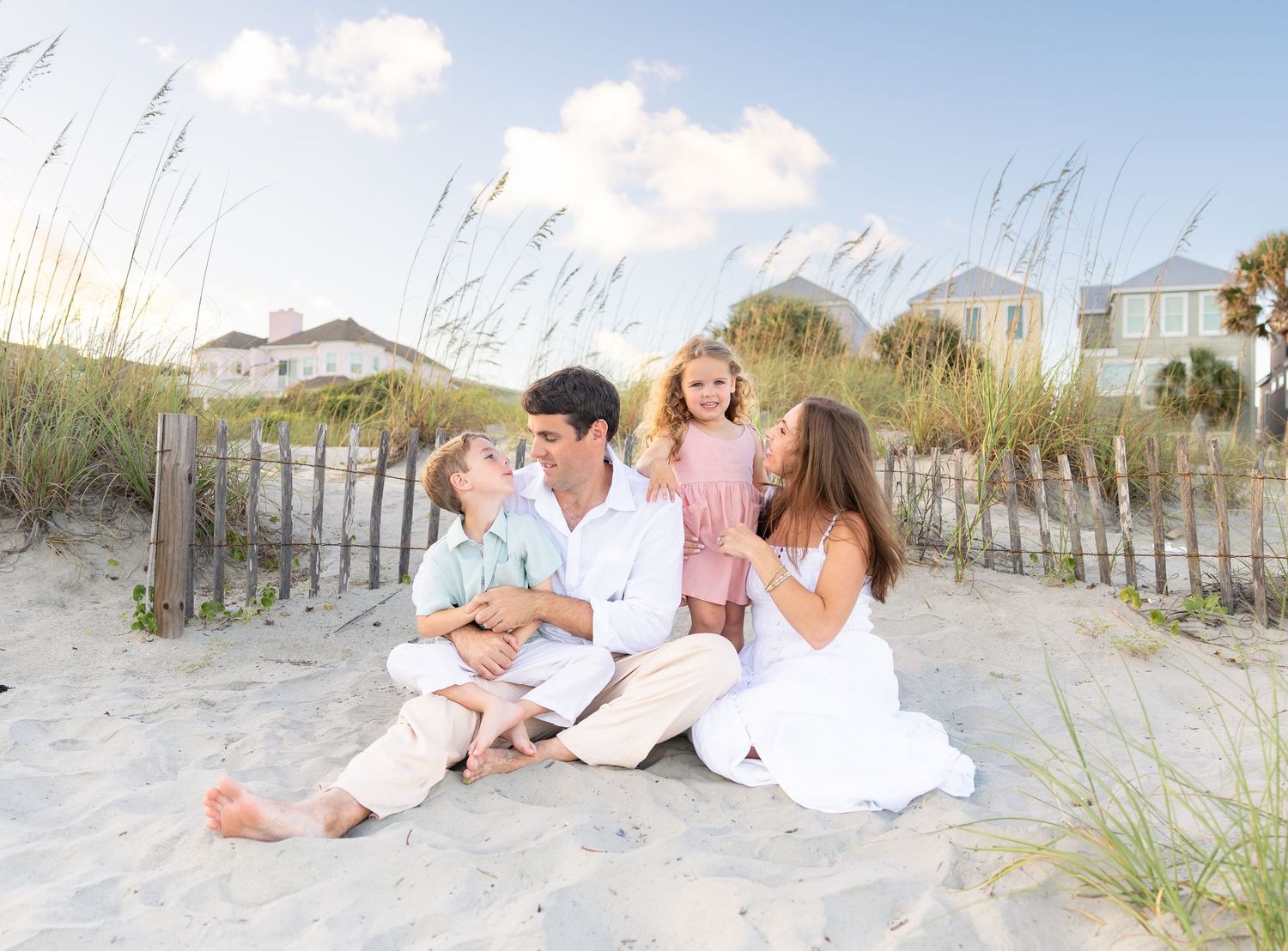 Family of four sitting together on sandy beach dunes near a wooden fence, enjoying a sunny day by the ocean.