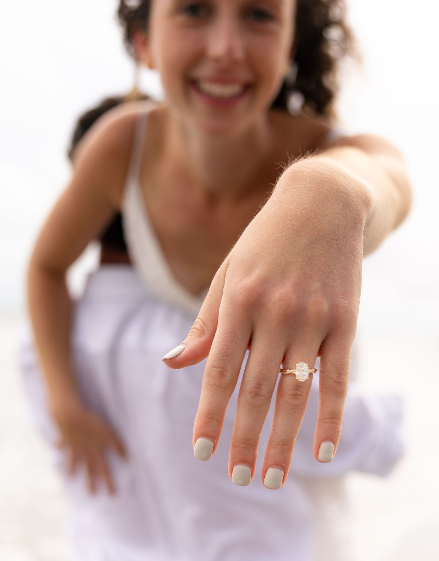 Woman smiling on beach displaying diamond engagement ring on outstretched hand with neutral manicure.