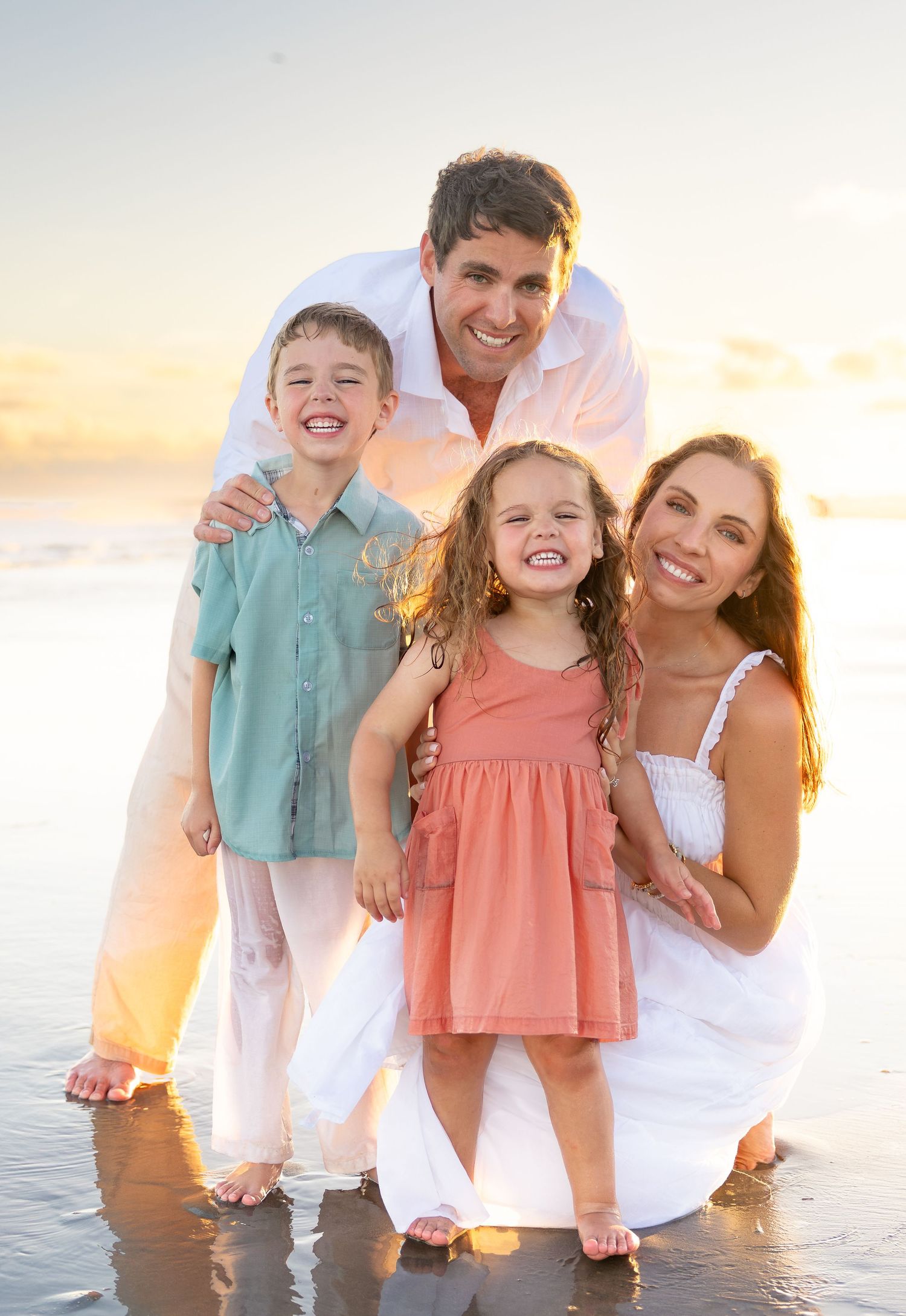 Happy family of four posing on a sunny beach at sunset, parents and two young children smiling in pastel outfits.