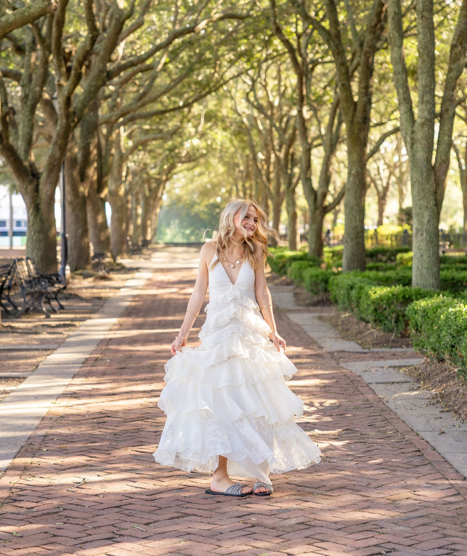 Woman in a white tiered ruffle dress walks down a brick path lined with live oak trees and hedges in a sunlit park.