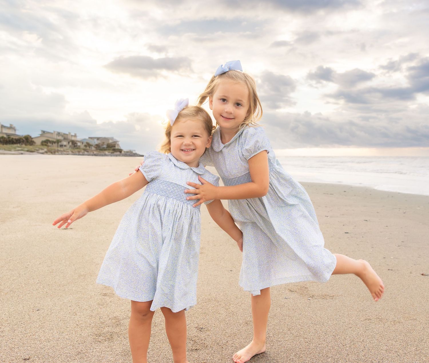Two young girls in matching light blue smocked dresses and hair bows laugh and play together on a sandy beach at sunset.