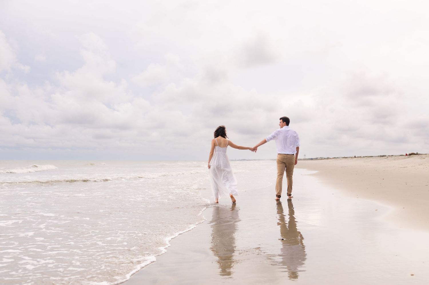 Couple holding hands walking along a wet sandy beach at the water's edge, the bride in a white dress under a cloudy sky.