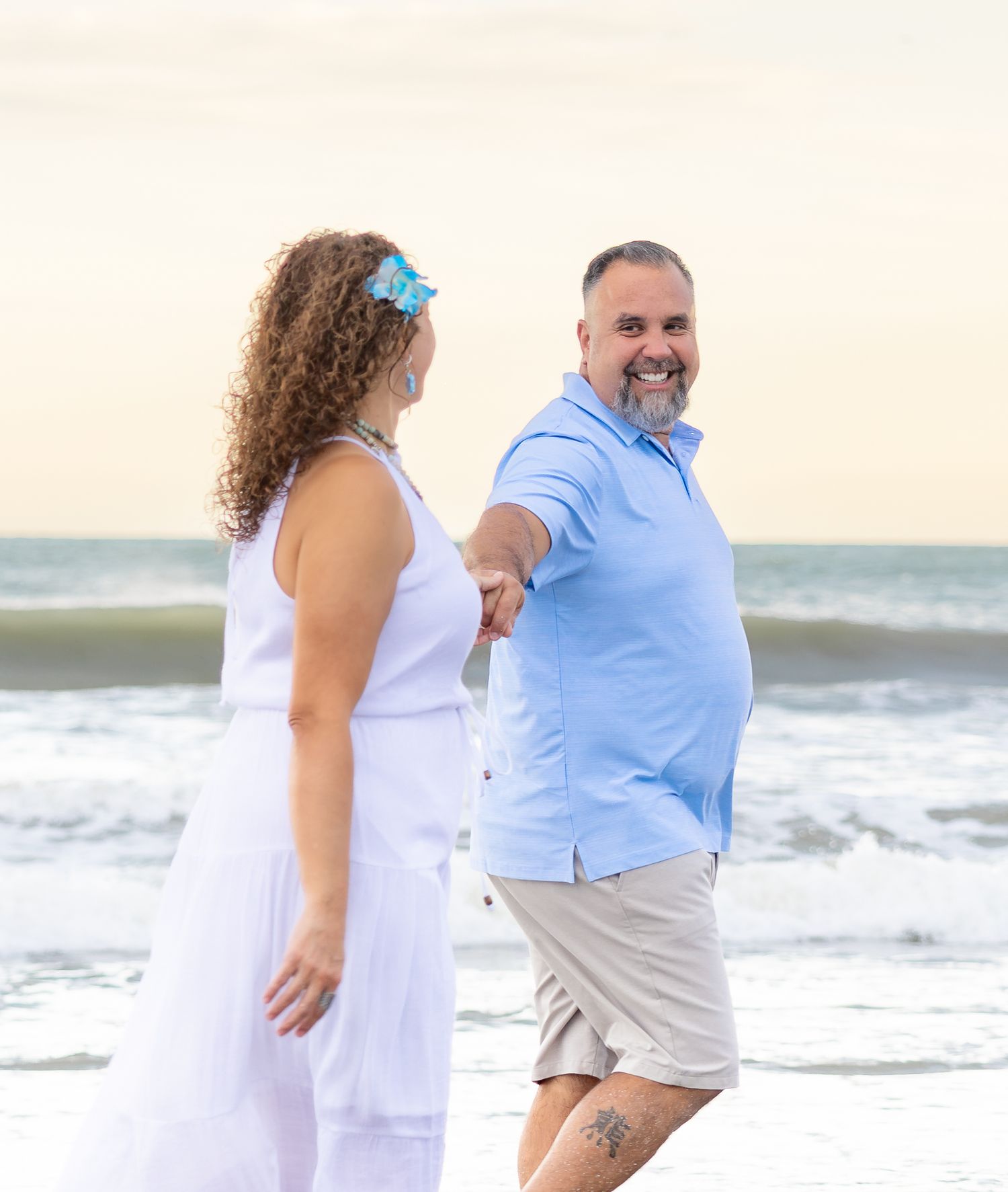 Smiling couple walking hand in hand along the beach at sunset, the woman in a white dress with a blue flower and the man in a blue shirt.