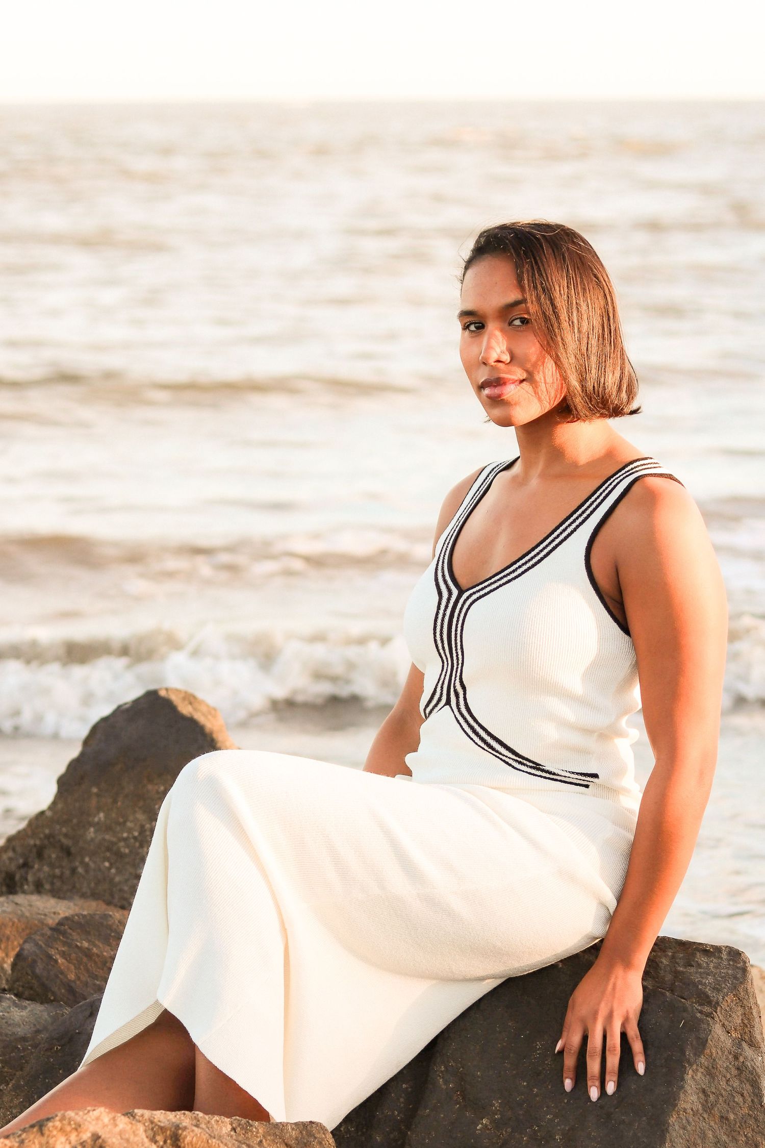 Woman in white dress with black trim sits on rocks by ocean waves at sunset.