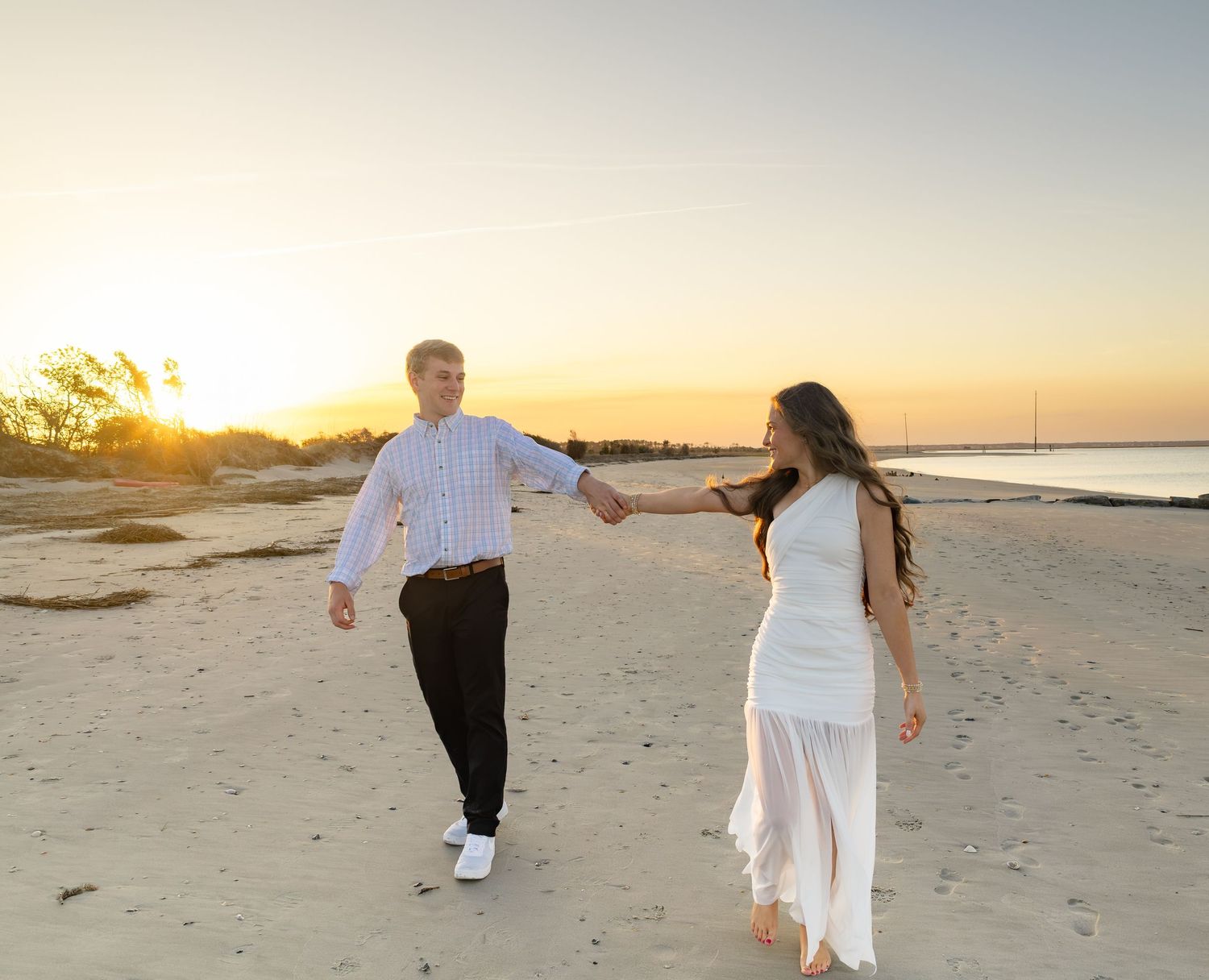 Couple holding hands and dancing on a sandy beach at sunset, woman in white dress, man in striped shirt.